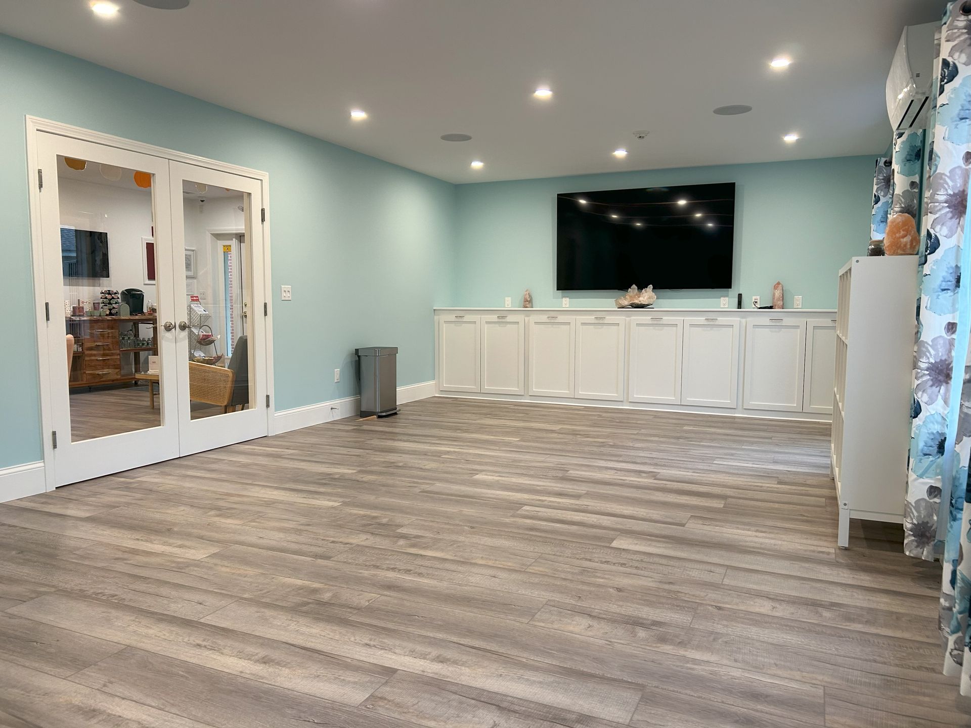Spacious, light-blue room with a large TV, white cabinets, French doors, and wood-look flooring.