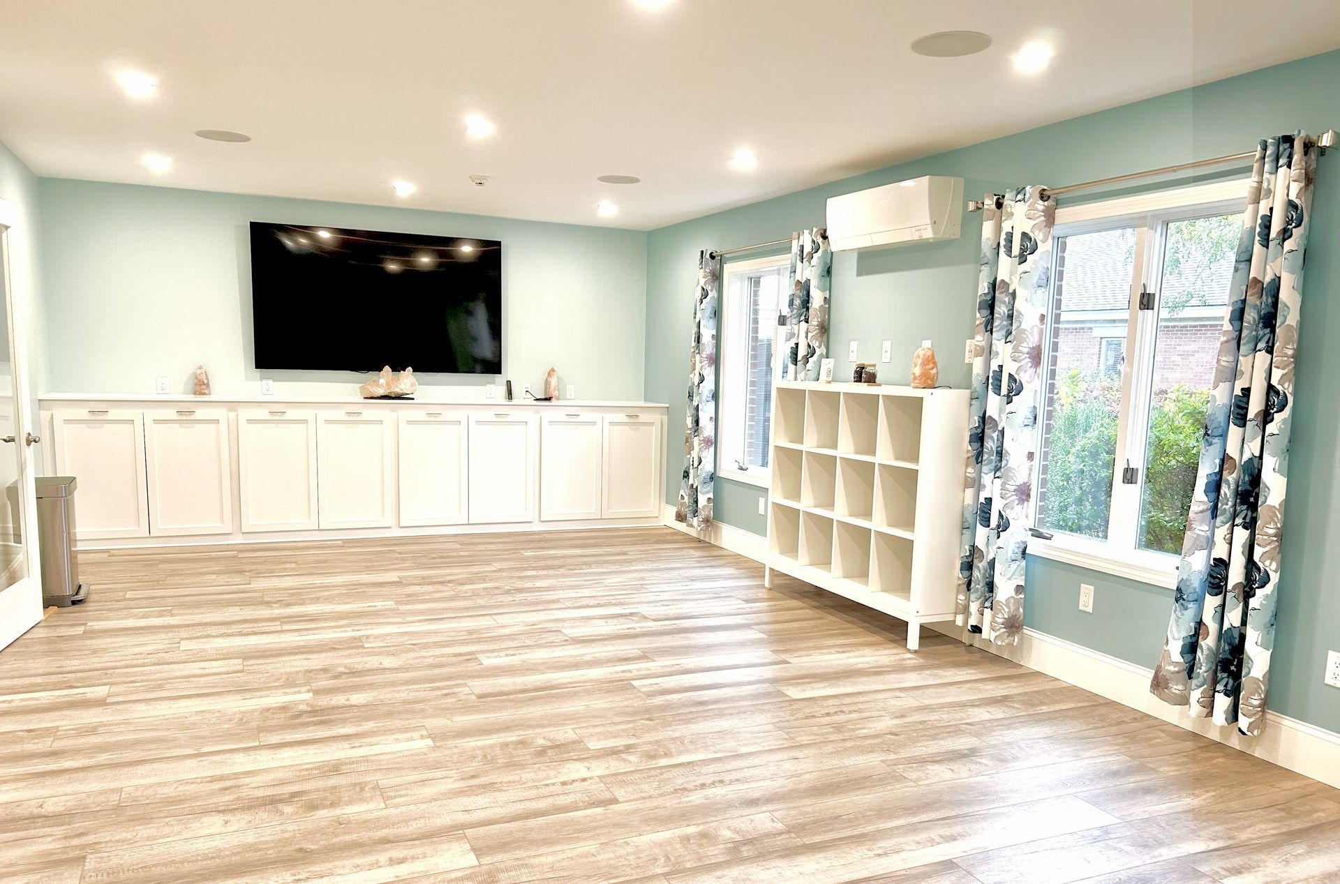 Spacious room with light blue walls, light wood floor, large TV, white cabinets, and white shelf unit near windows with patterned curtains.