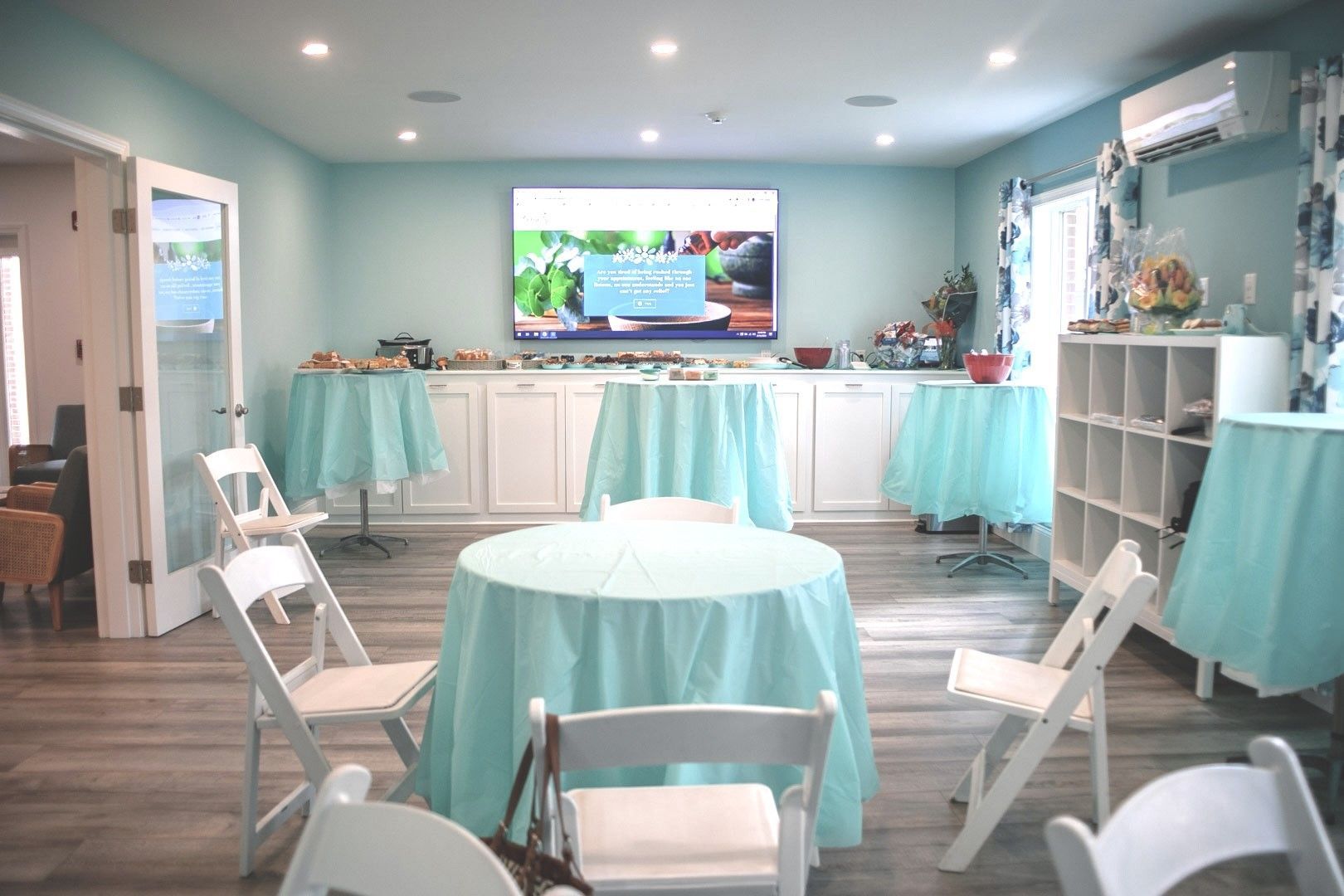 A room with tables draped in teal, white folding chairs, and food displays. A TV screen shows a food image.