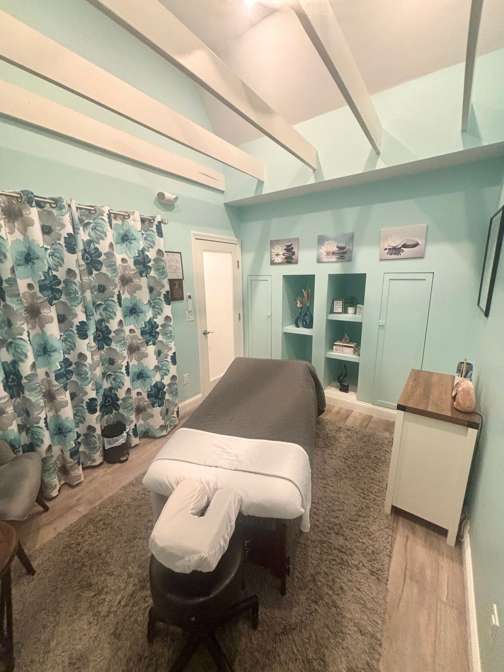 Massage room: massage table, teal walls, floral curtains, and beige rug.