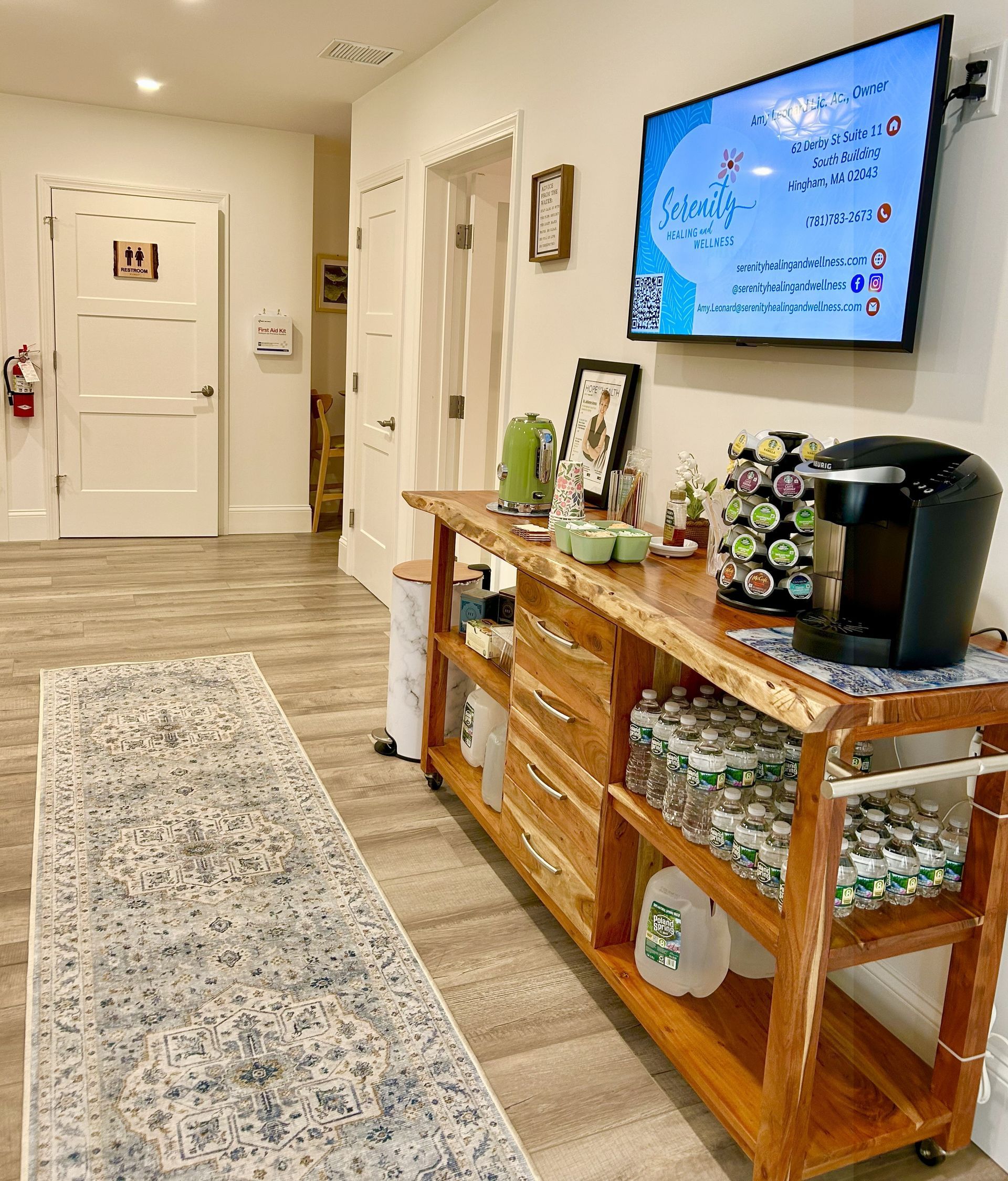 Hallway with a wooden coffee station, TV, bench, and doors. The station has a coffee maker, snacks, and bottled water.