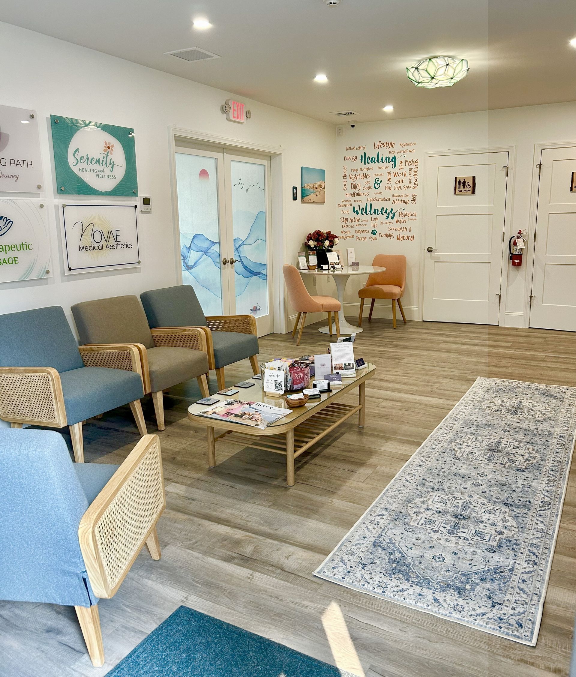 Waiting room with light wood floors, blue armchairs, and a coffee table. A rug leads to a doorway with the exit sign.