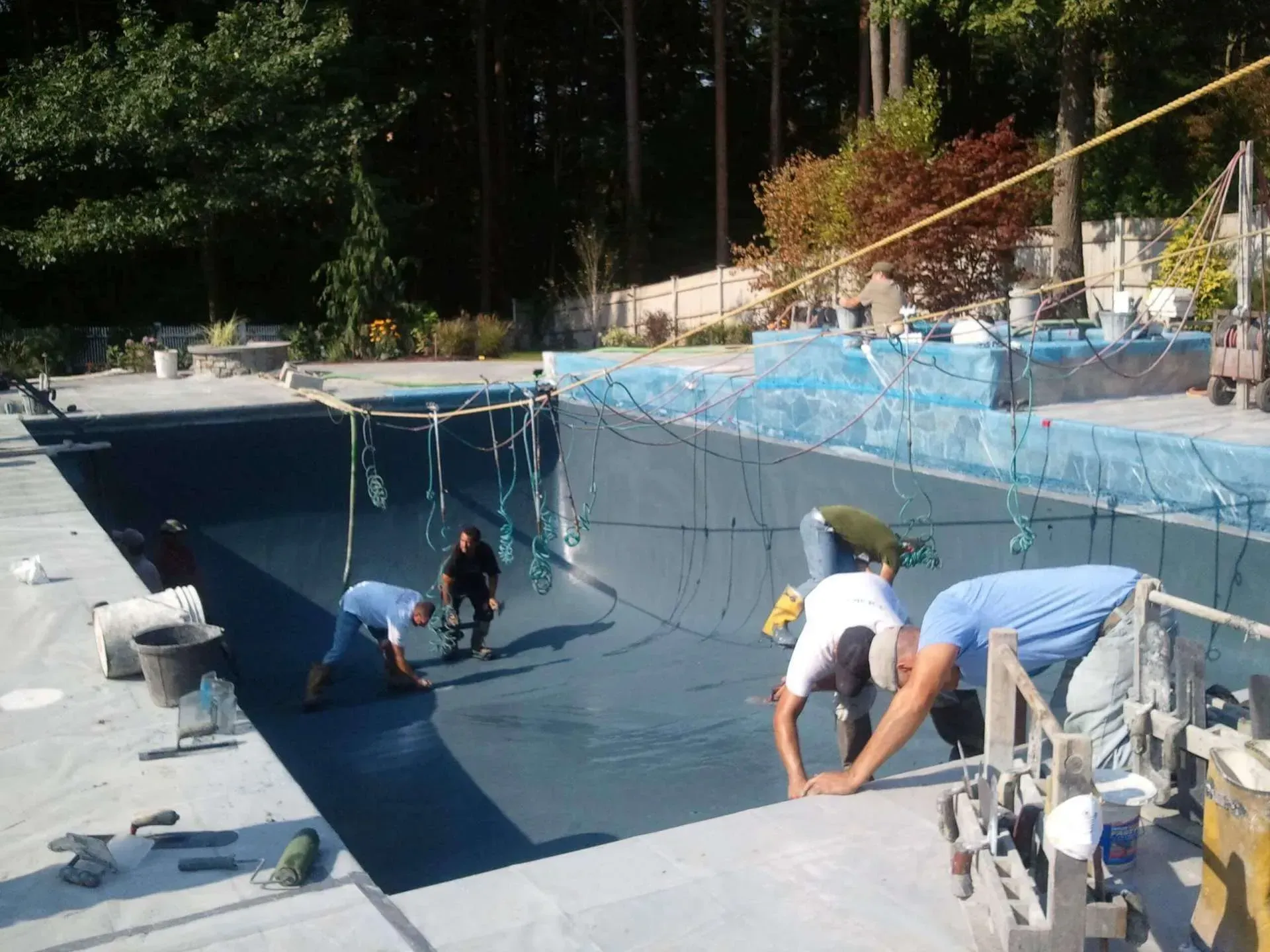 Workers installing a pool liner. They're bent over inside the pool. Light blue liner, concrete edges, trees in background.