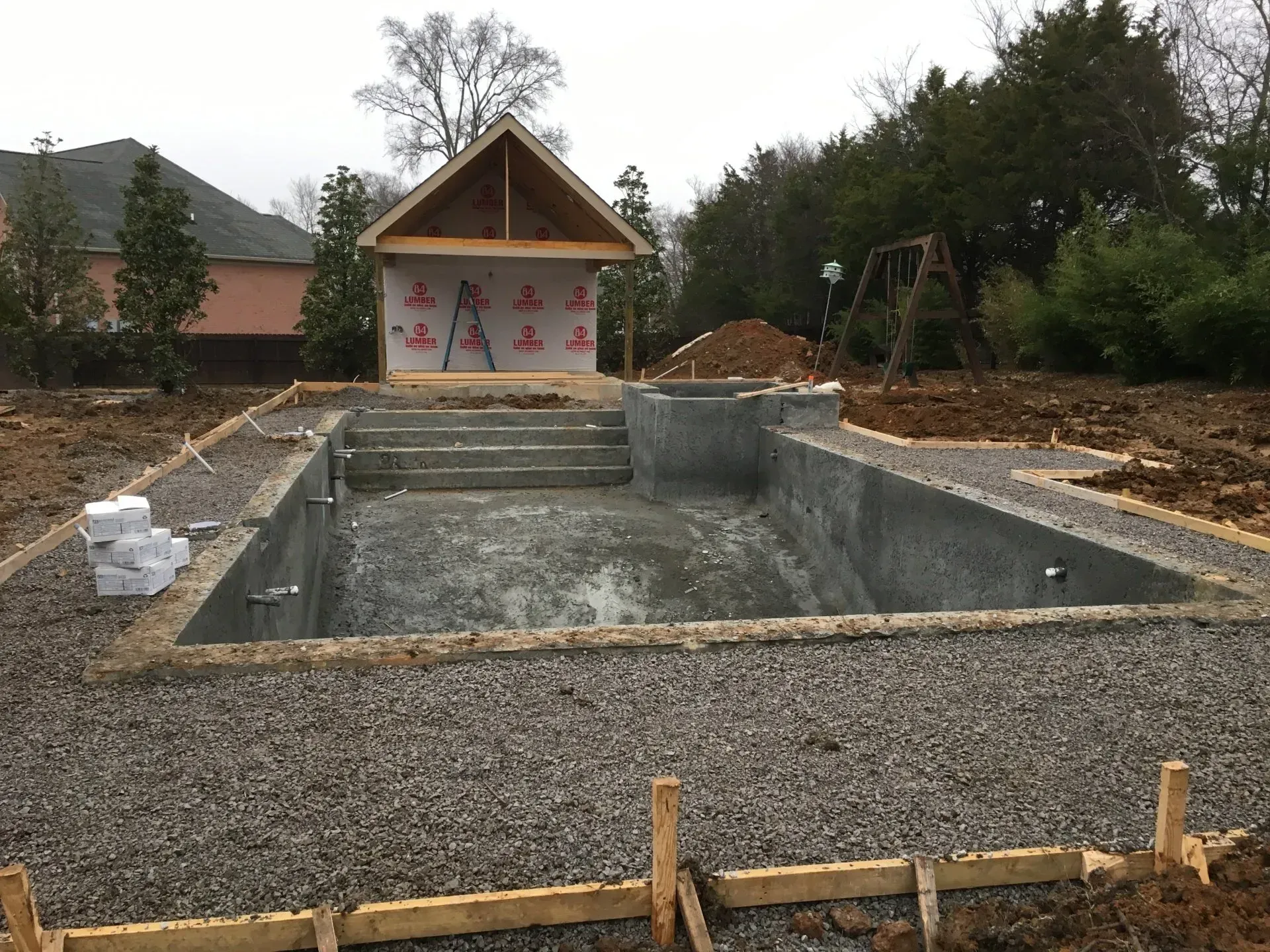 Pool construction site: concrete pool shell with steps, unfinished shed, gravel surround.