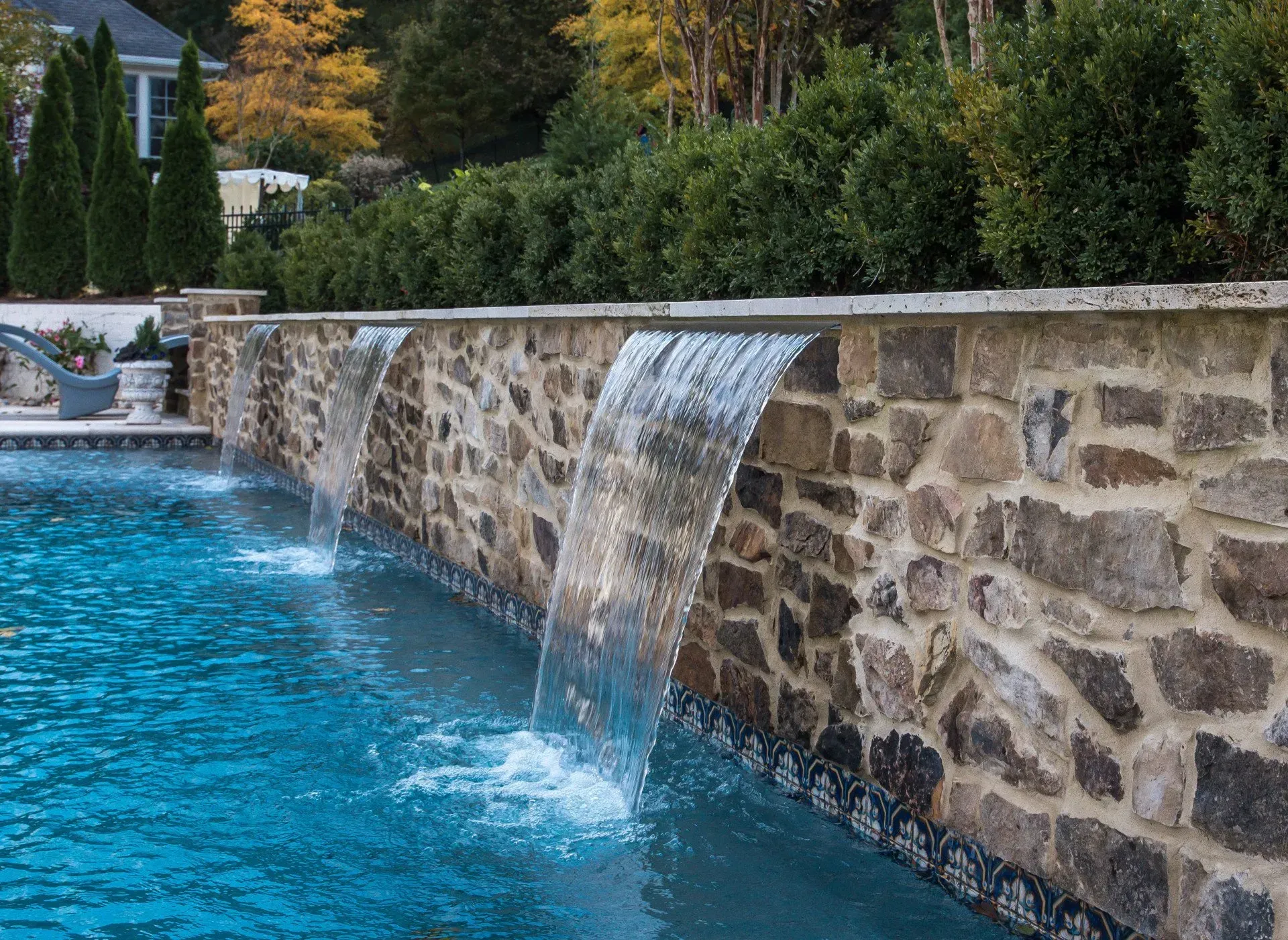 Pool with stone wall waterfall feature, blue water, green shrubbery in background.