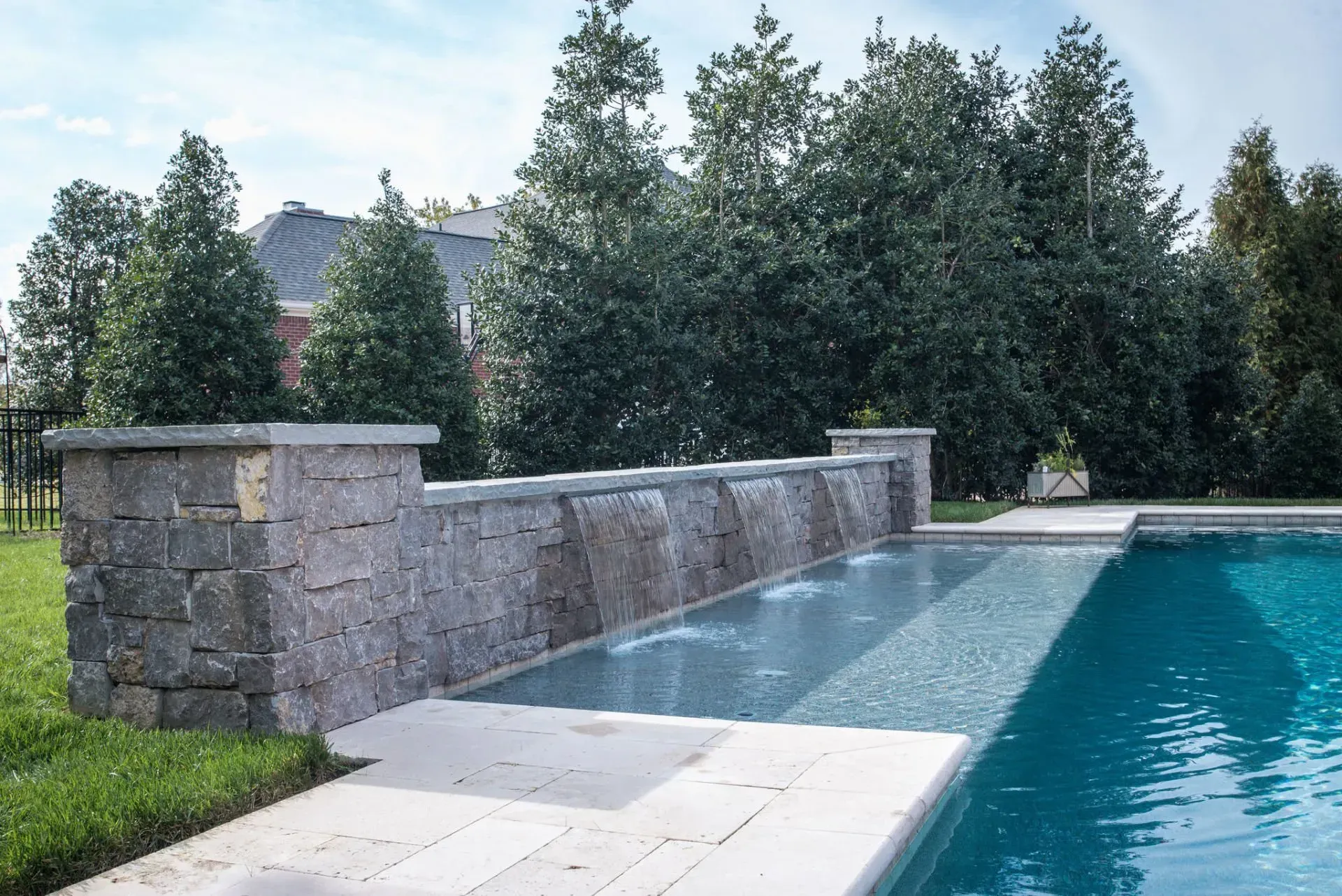 A stone wall with water features flowing into a blue pool, surrounded by trees and green grass.