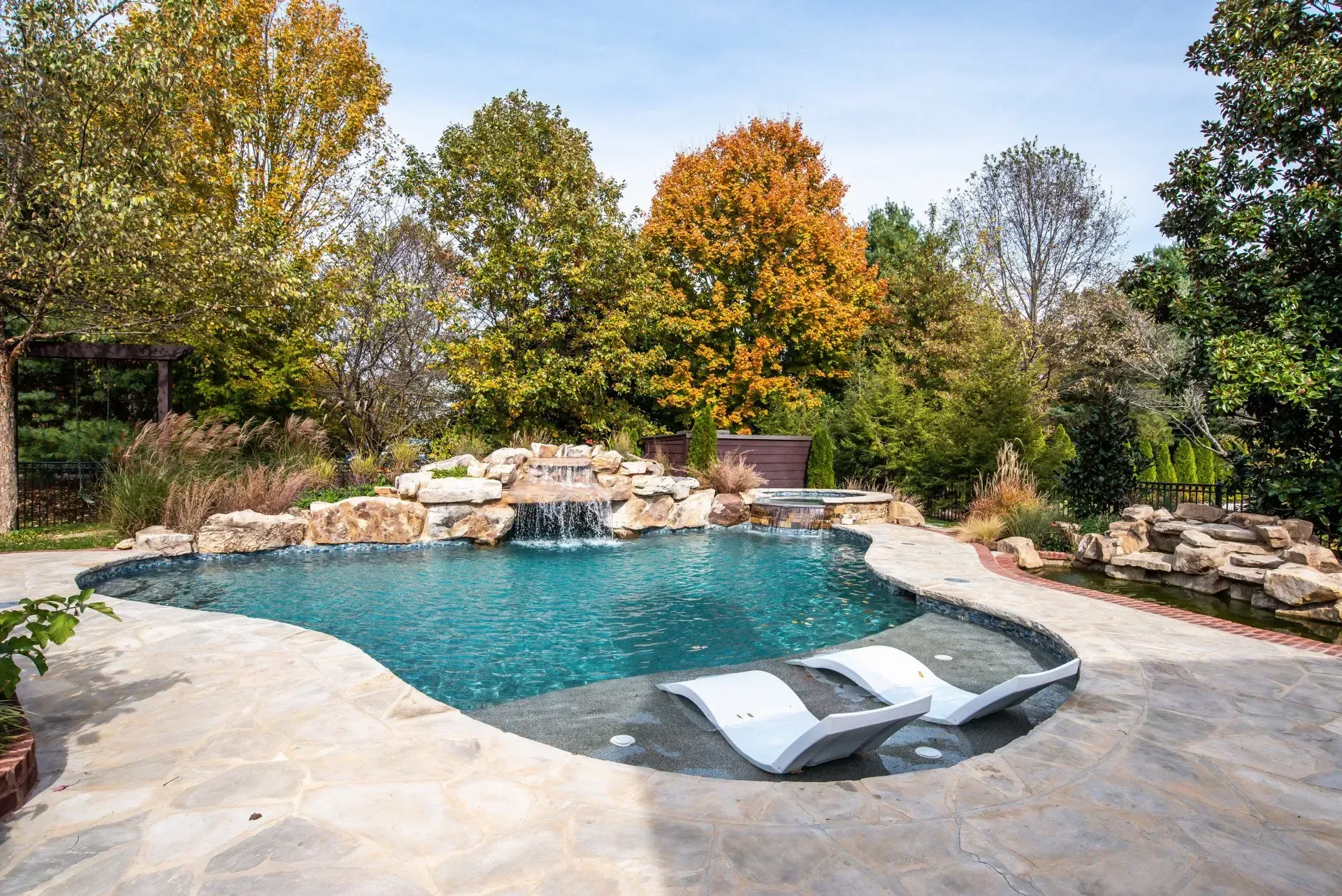 Pool with waterfalls, stone patio, and lounge chairs surrounded by trees with fall foliage.