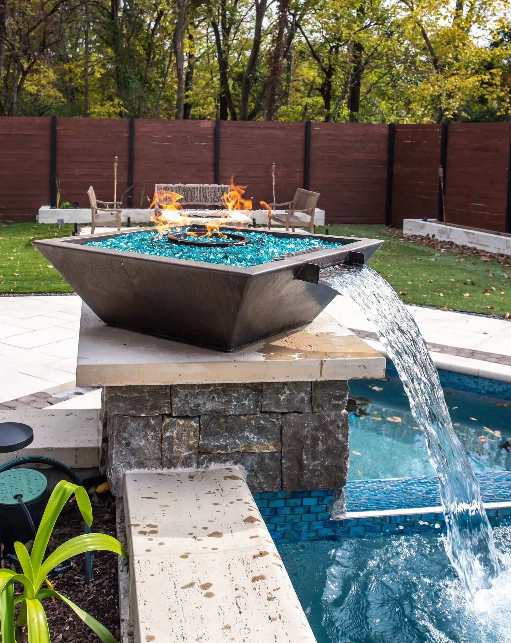 Fire pit with blue glass and flames over pool with water flowing. Brown fence in background.