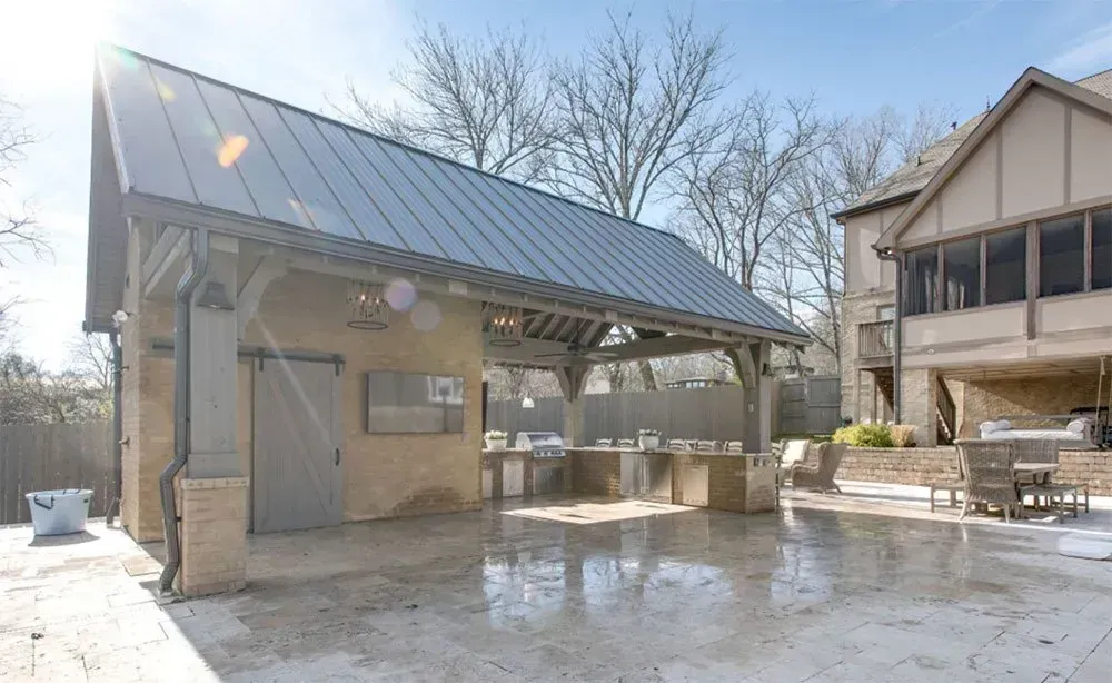 Outdoor kitchen with gray metal roof, brick, concrete patio, and adjacent house.
