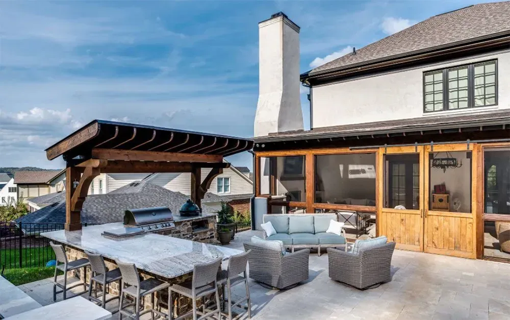 Outdoor patio with a kitchen area, seating, and a screened-in porch, adjacent to a two-story house.