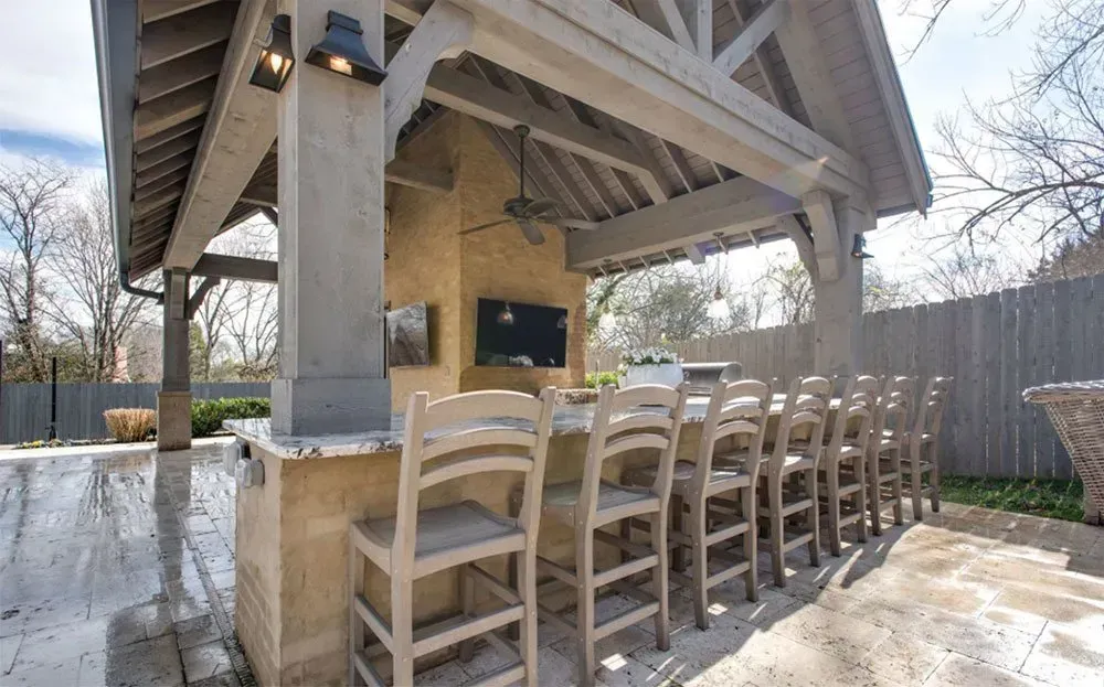 Outdoor kitchen with a bar, seating, and a TV, all under a covered structure with stone and wood elements.