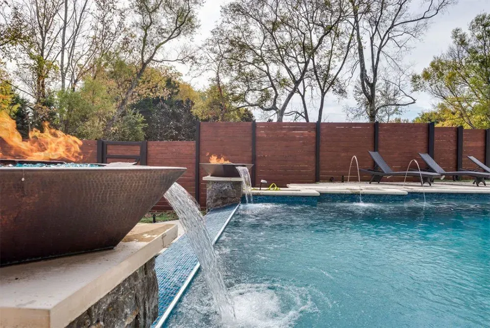 A pool with a fire feature, water fountain, and lounge chairs next to a wooden fence.