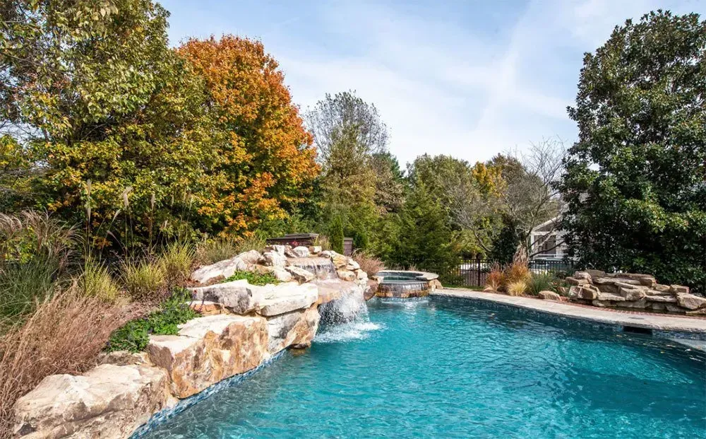 Swimming pool with waterfall feature, surrounded by trees with fall foliage.