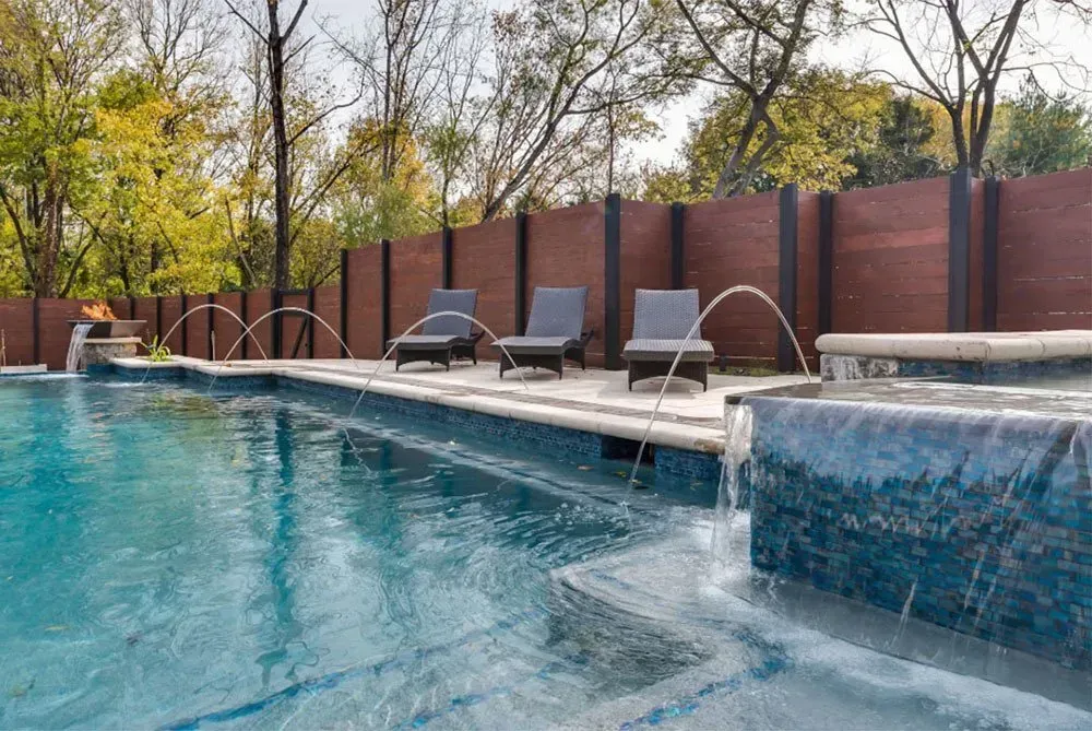 A luxurious pool with fountains and lounge chairs, a wooden fence, and trees.