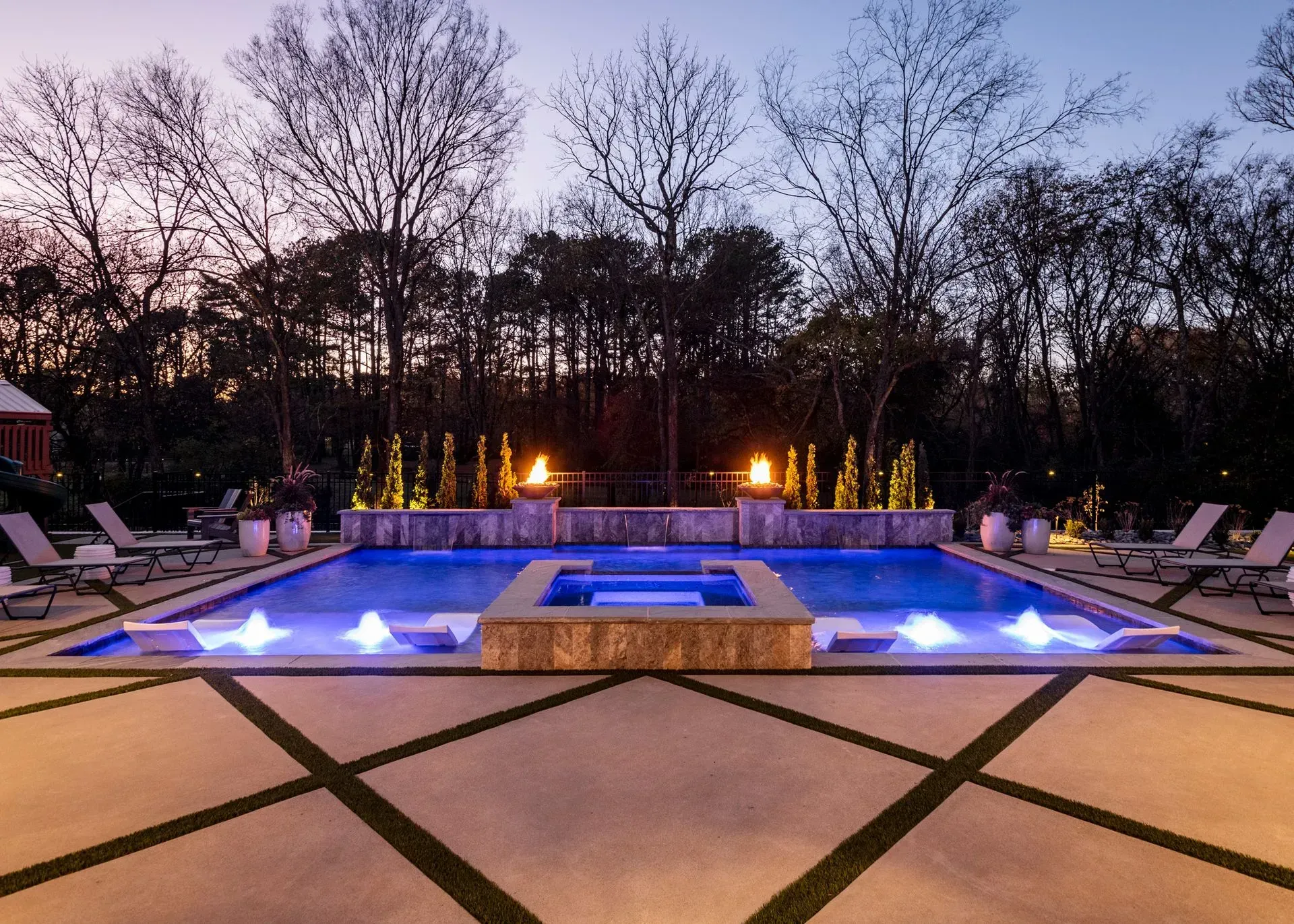 Luxury backyard pool at dusk with fire features, fountains, and lounge chairs.