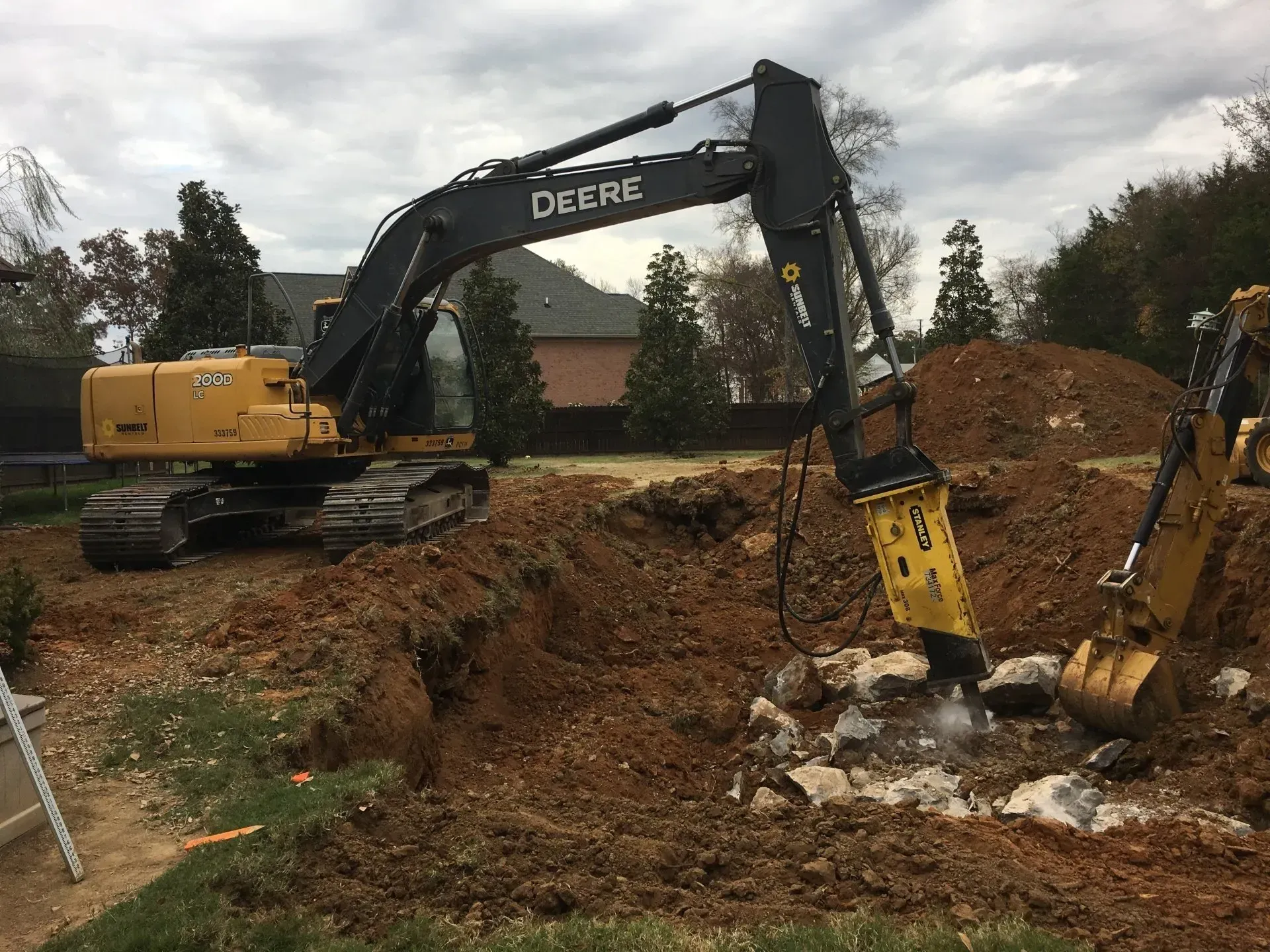A John Deere excavator uses a jackhammer attachment to break apart rocks in a dirt excavation site.