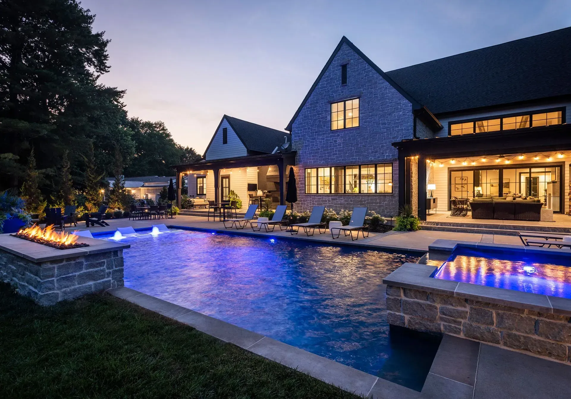 Luxury home exterior at dusk, lit pool with fire features, stone facade, dark roof.