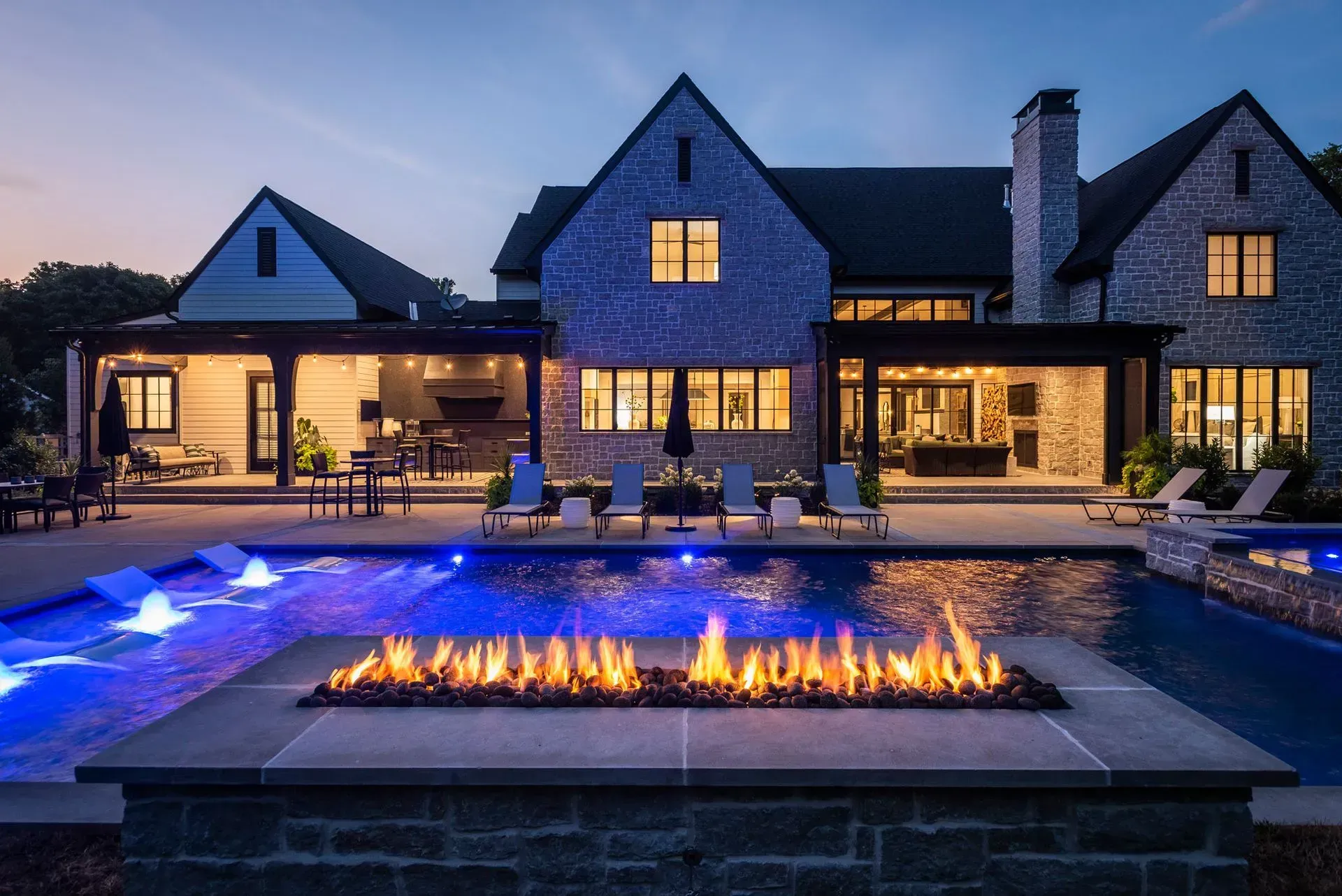 Luxury home with pool at dusk; fire pit, blue pool lights, and lounge chairs.