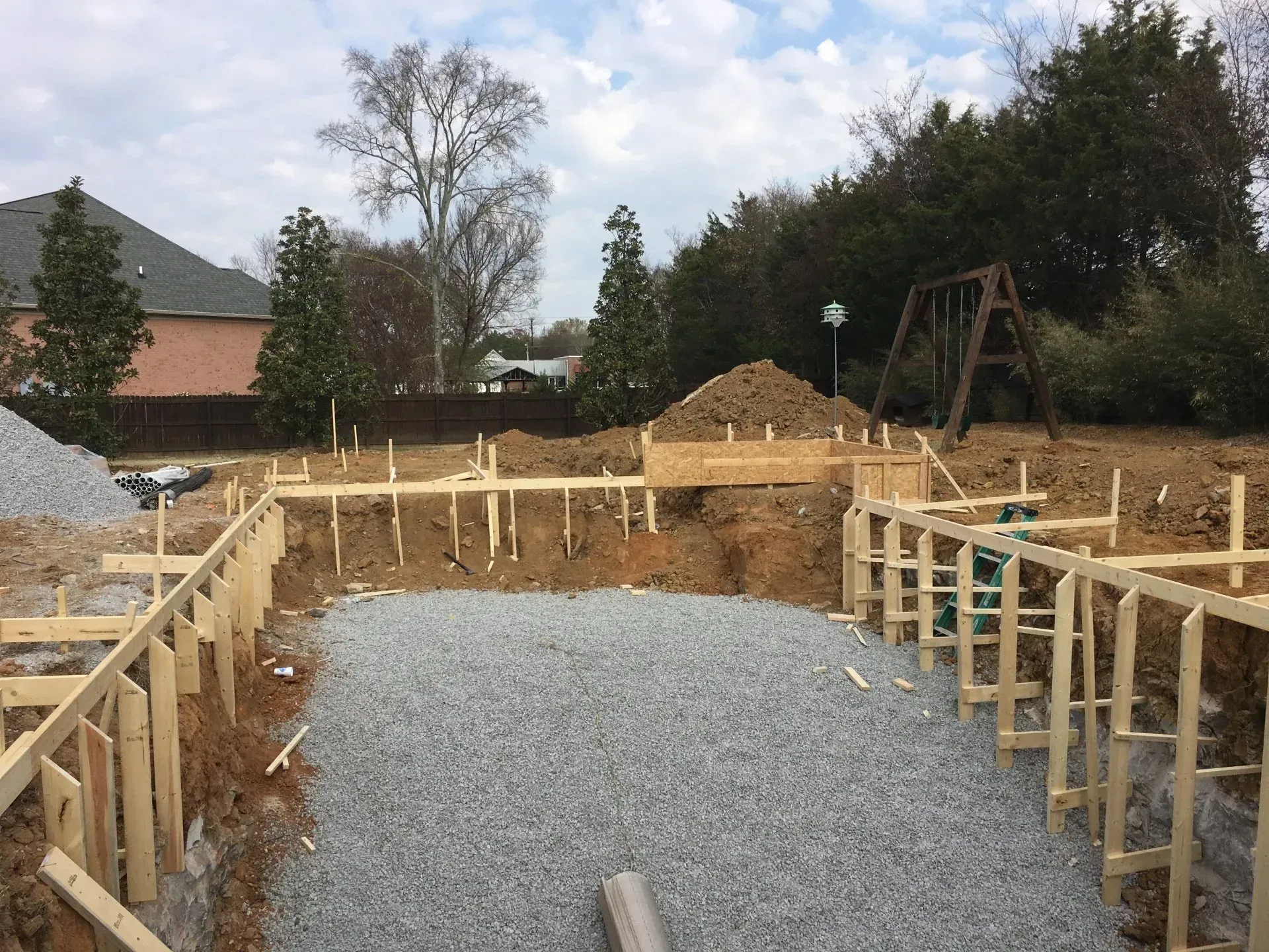 Construction site: wooden forms outline a gravel-filled foundation; soil piles and trees in background.