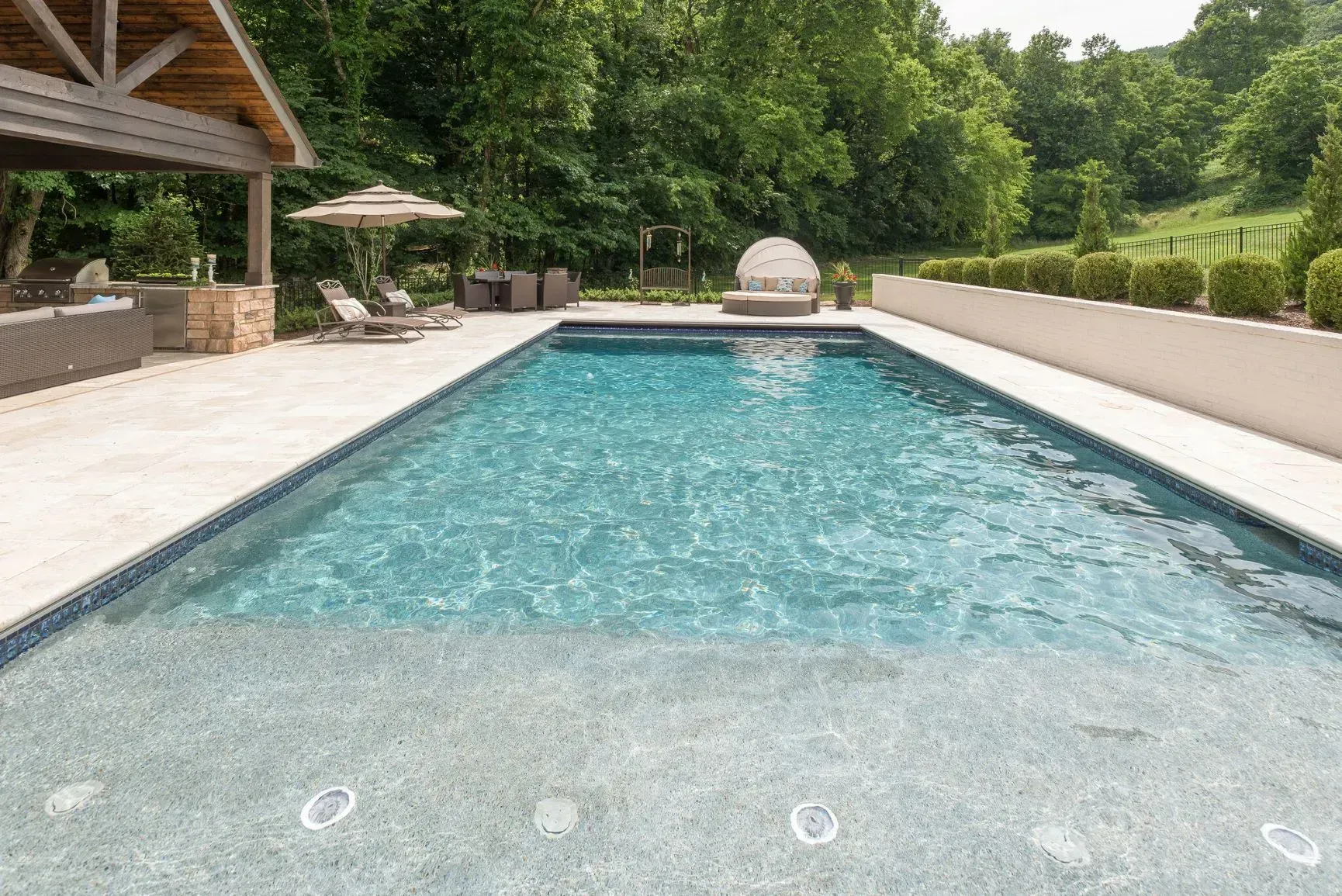 Rectangular swimming pool with blue water, patio, lounge chairs, and green trees.