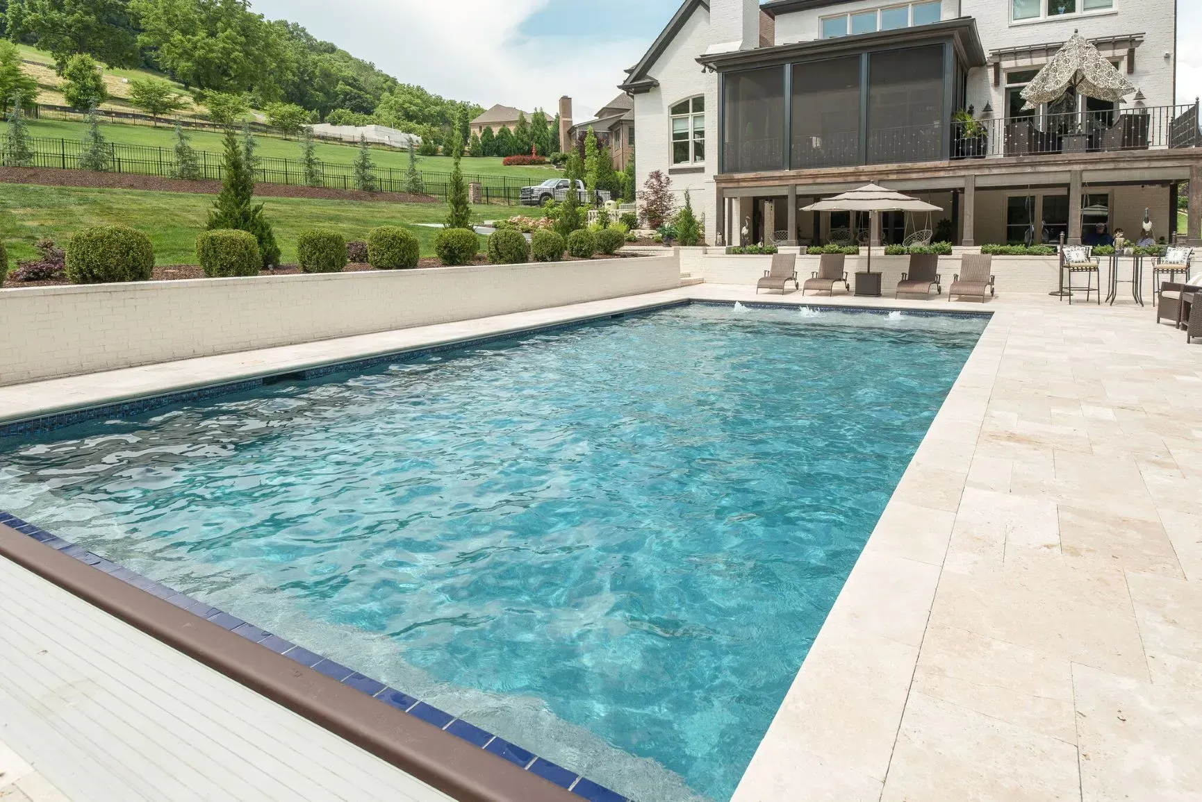 Swimming pool surrounded by a stone patio, with lounge chairs and a large house in the background.