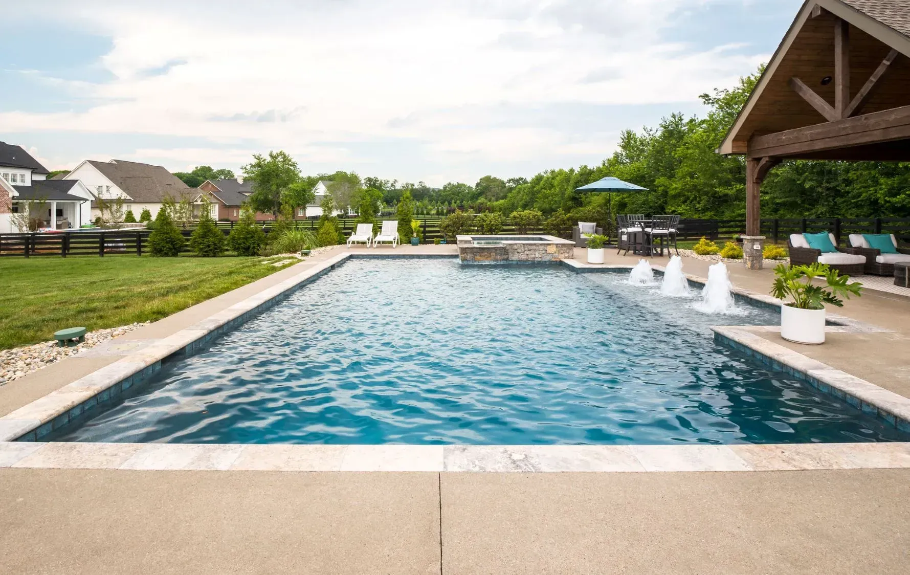 Swimming pool with fountains, a patio, and a view of houses and trees.
