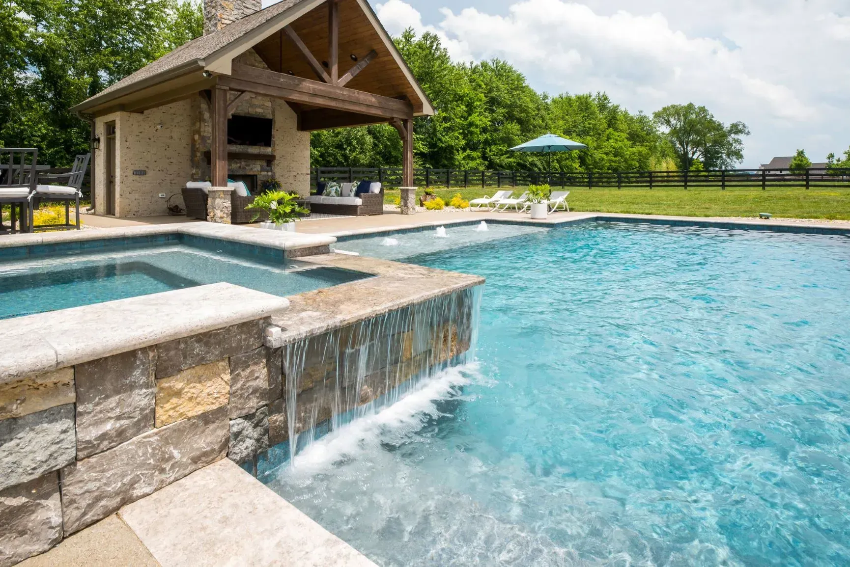 Pool with waterfall feature, hot tub, and covered outdoor kitchen. Blue water, stone construction, sunny day.