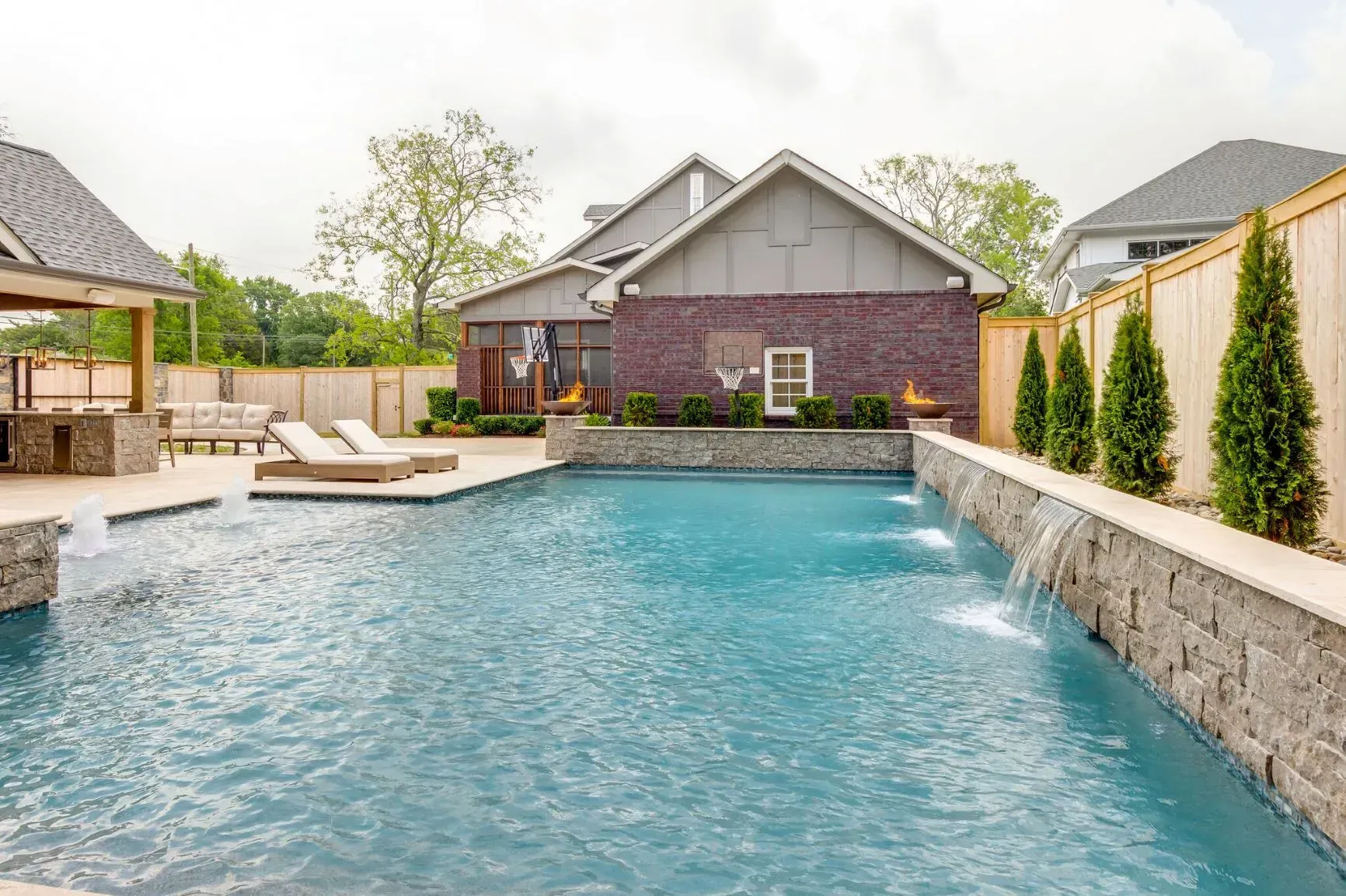 Swimming pool in a backyard with water fountains, brick house, lounge chairs, and a gazebo.