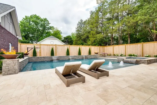 A luxury pool with two chaise lounges, a water feature, and a wooden fence.