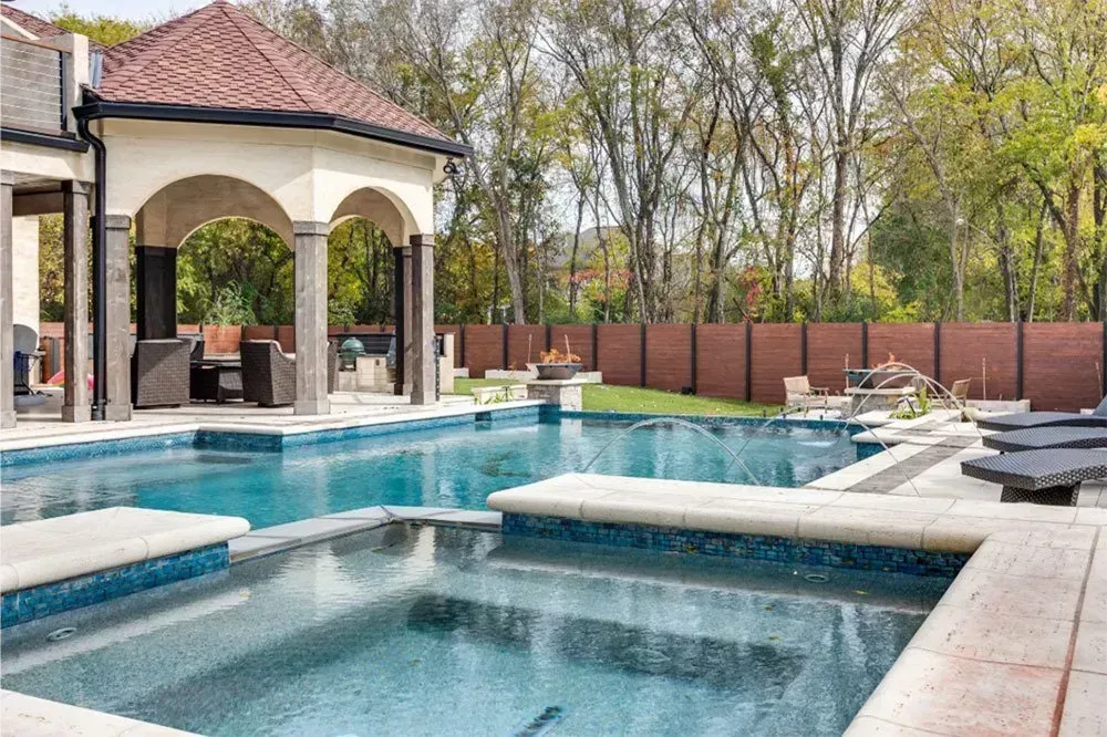 Luxurious backyard with pool and spa, tan patio, and arbor; trees in background.