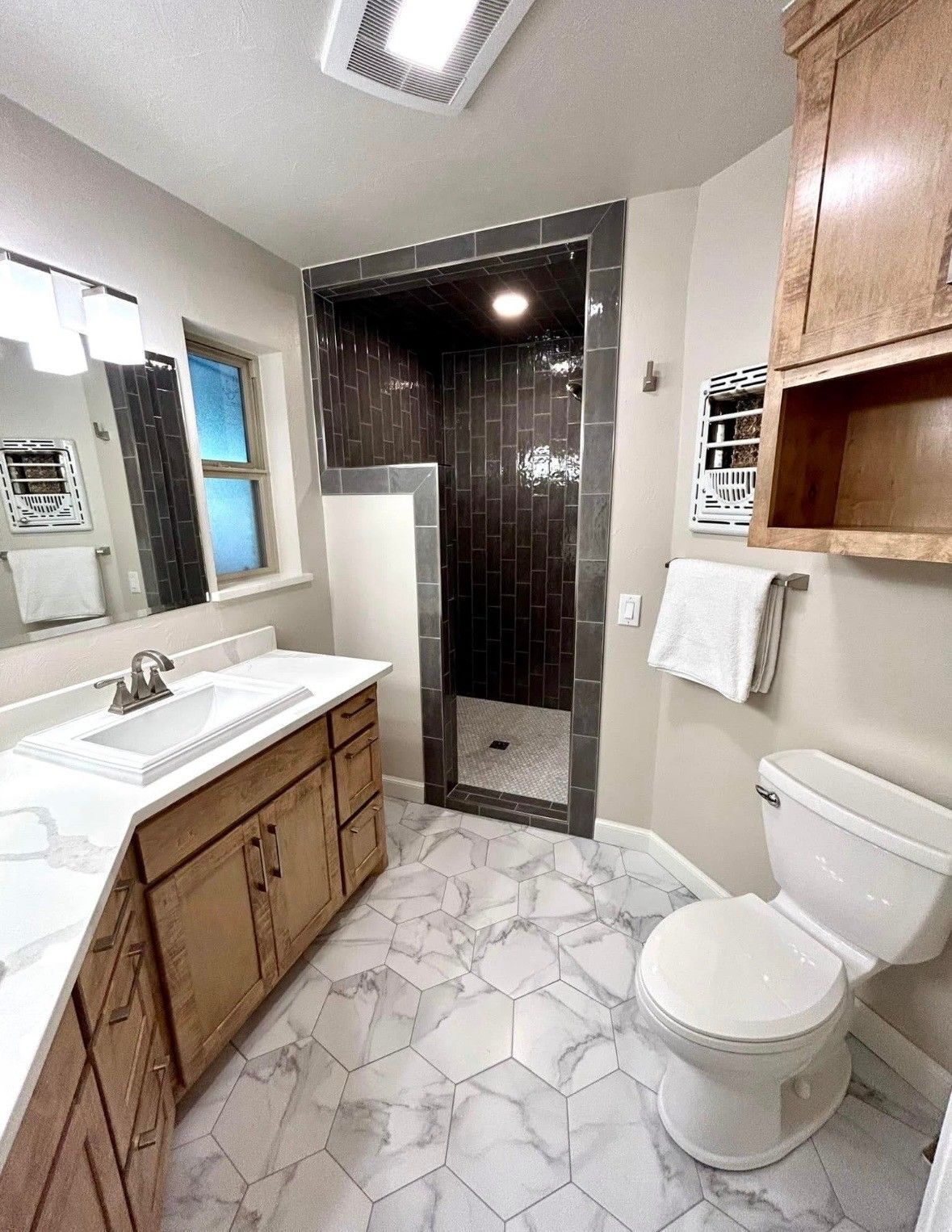 Bathroom with white and gray marble tile, wood vanity and cabinets, and a dark tile shower.