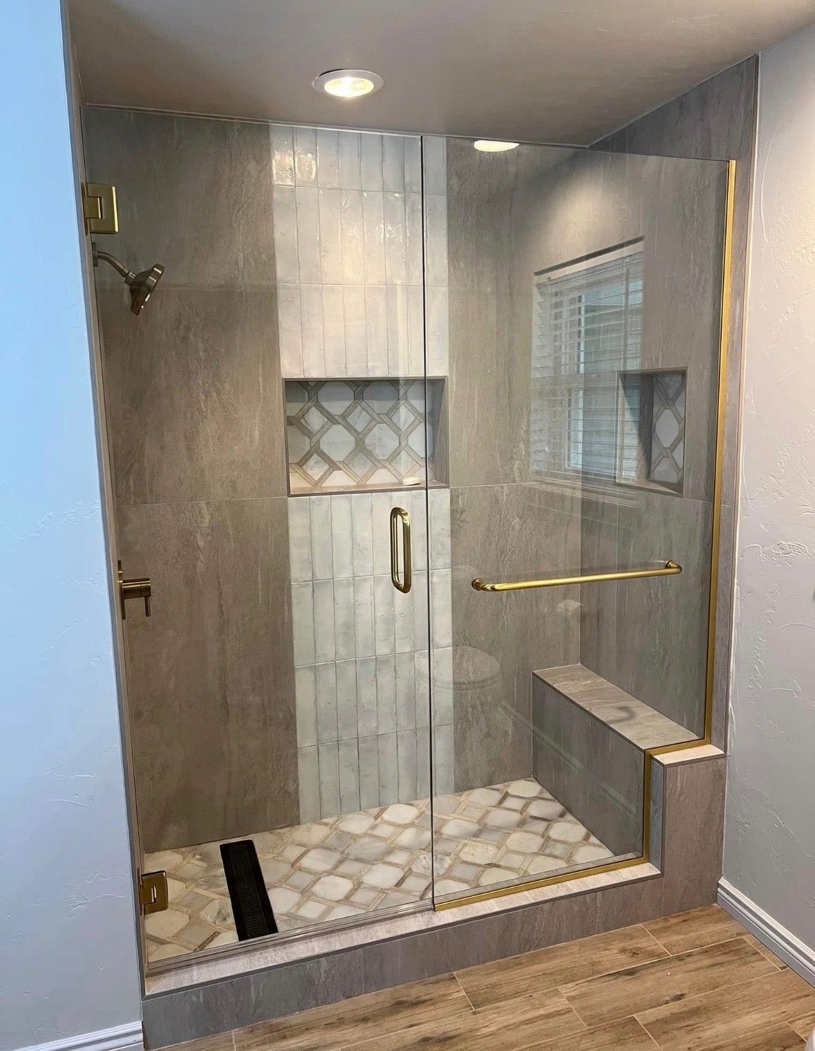 Modern gray tiled shower with gold fixtures and glass door, includes a bench and niche.