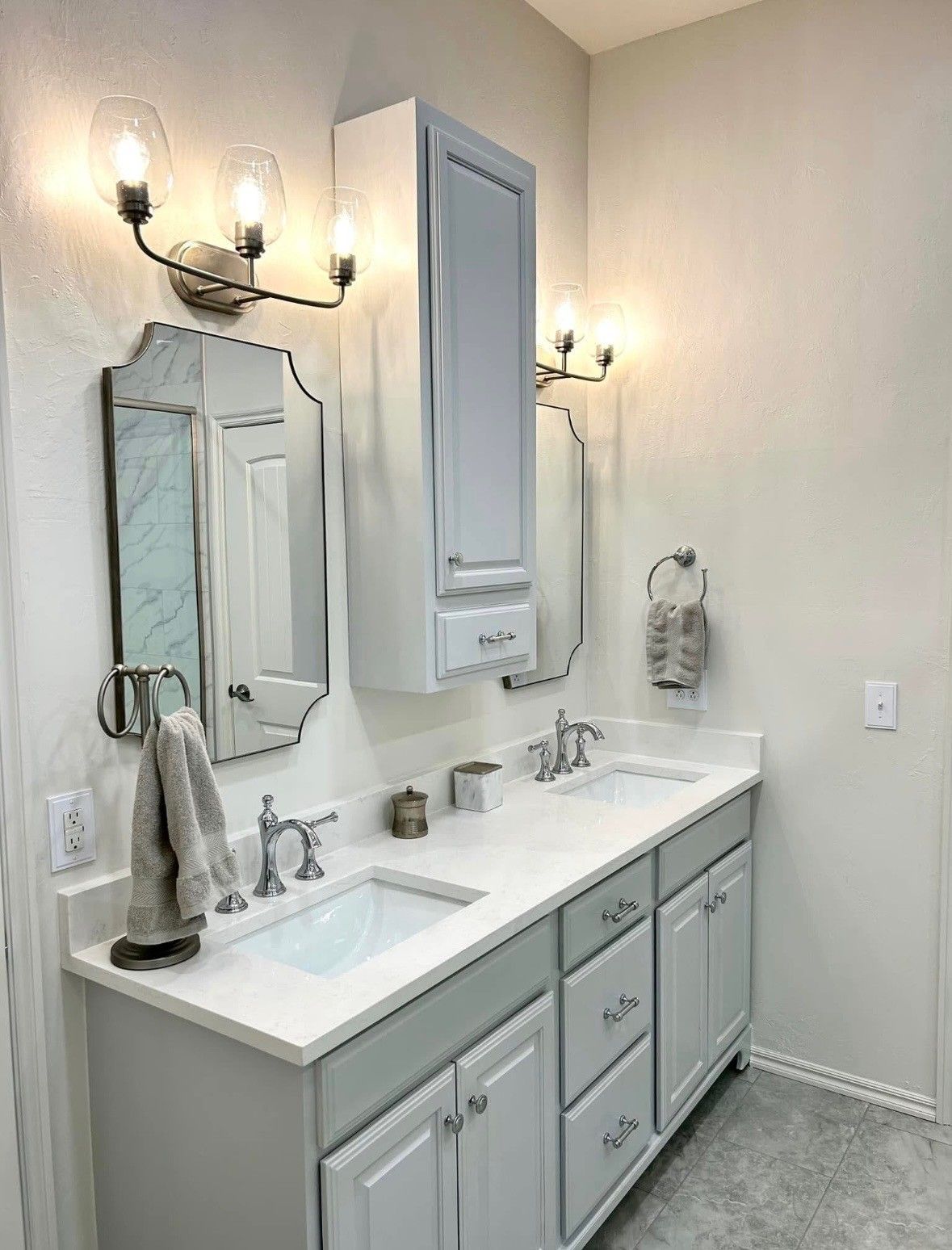A bathroom with double sinks, gray and white cabinets, mirrors, and a tall cabinet.