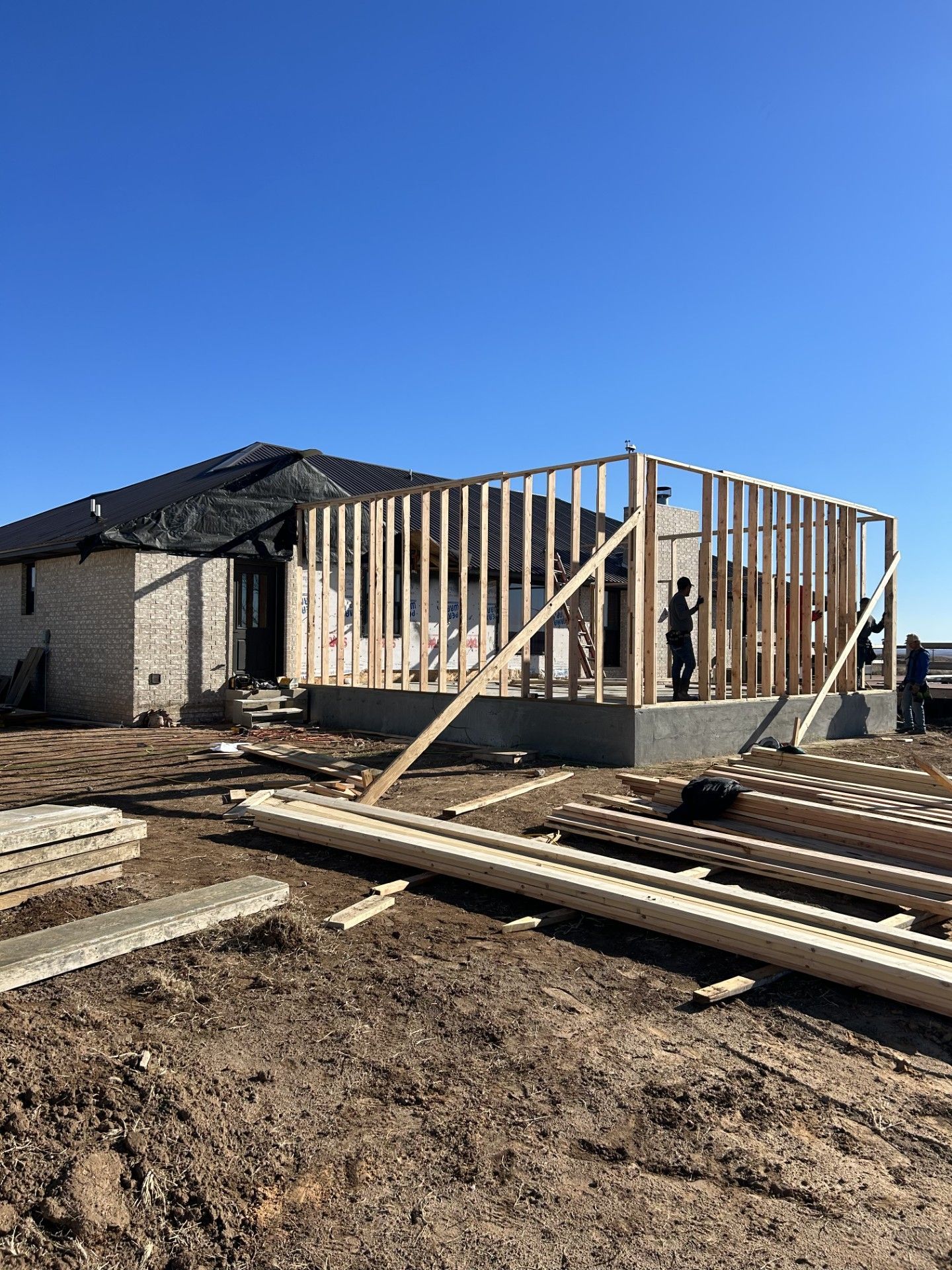Construction site with a partially framed house; workers erect walls on a concrete foundation under a clear blue sky.