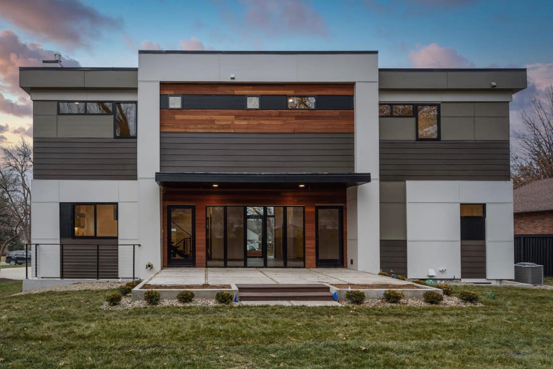 Modern two-story house with dark siding and wood accents, glass doors, and a concrete patio.