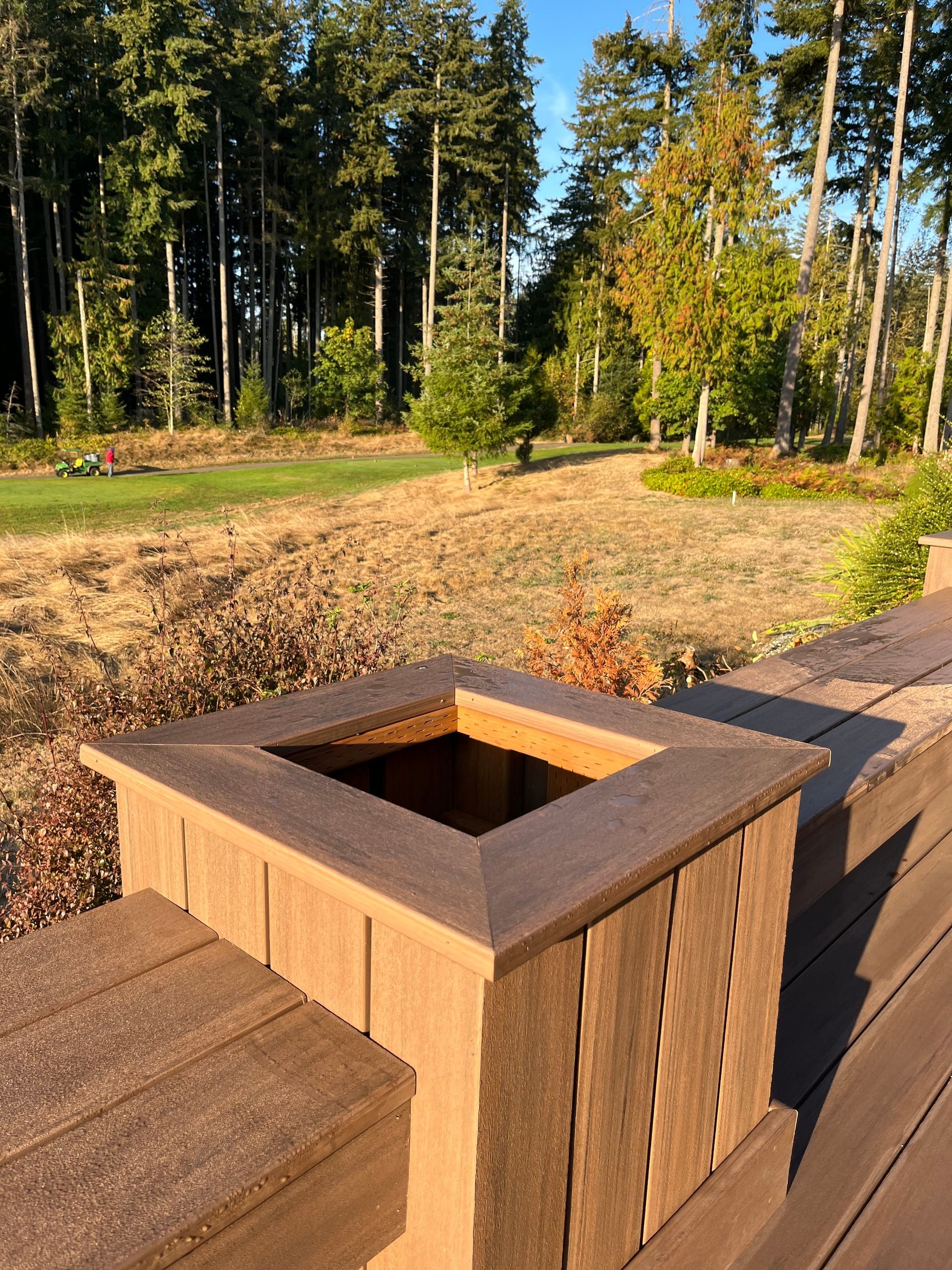 A wooden planter is sitting on a wooden deck with trees in the background.