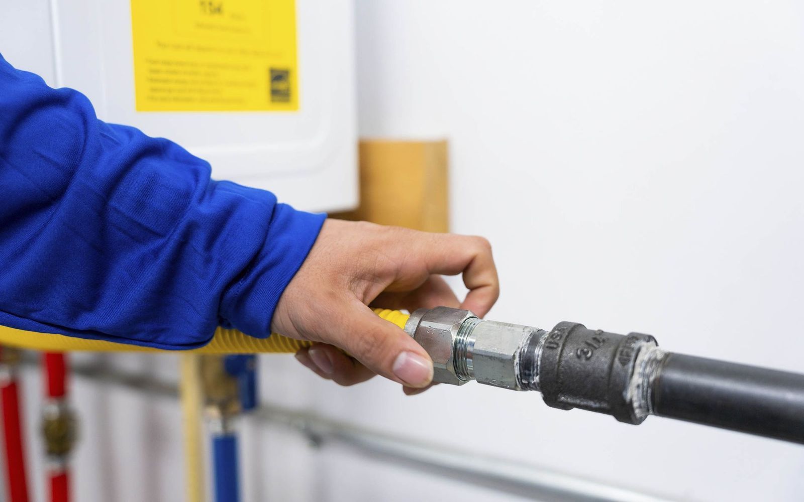 A person in blue overalls connects a yellow gas line to a black pipe near a water heater.