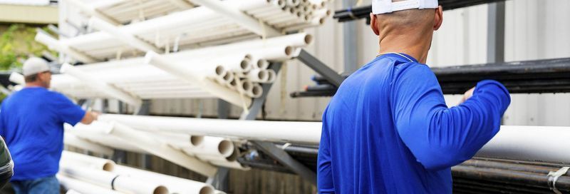 Two workers in blue shirts load pipes from a rack.