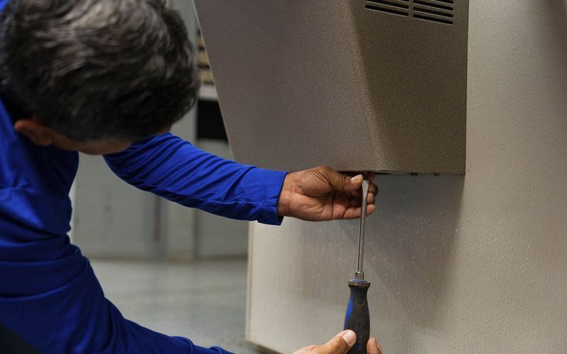 Man in blue shirt uses a screwdriver to work on a beige-colored wall-mounted object.
