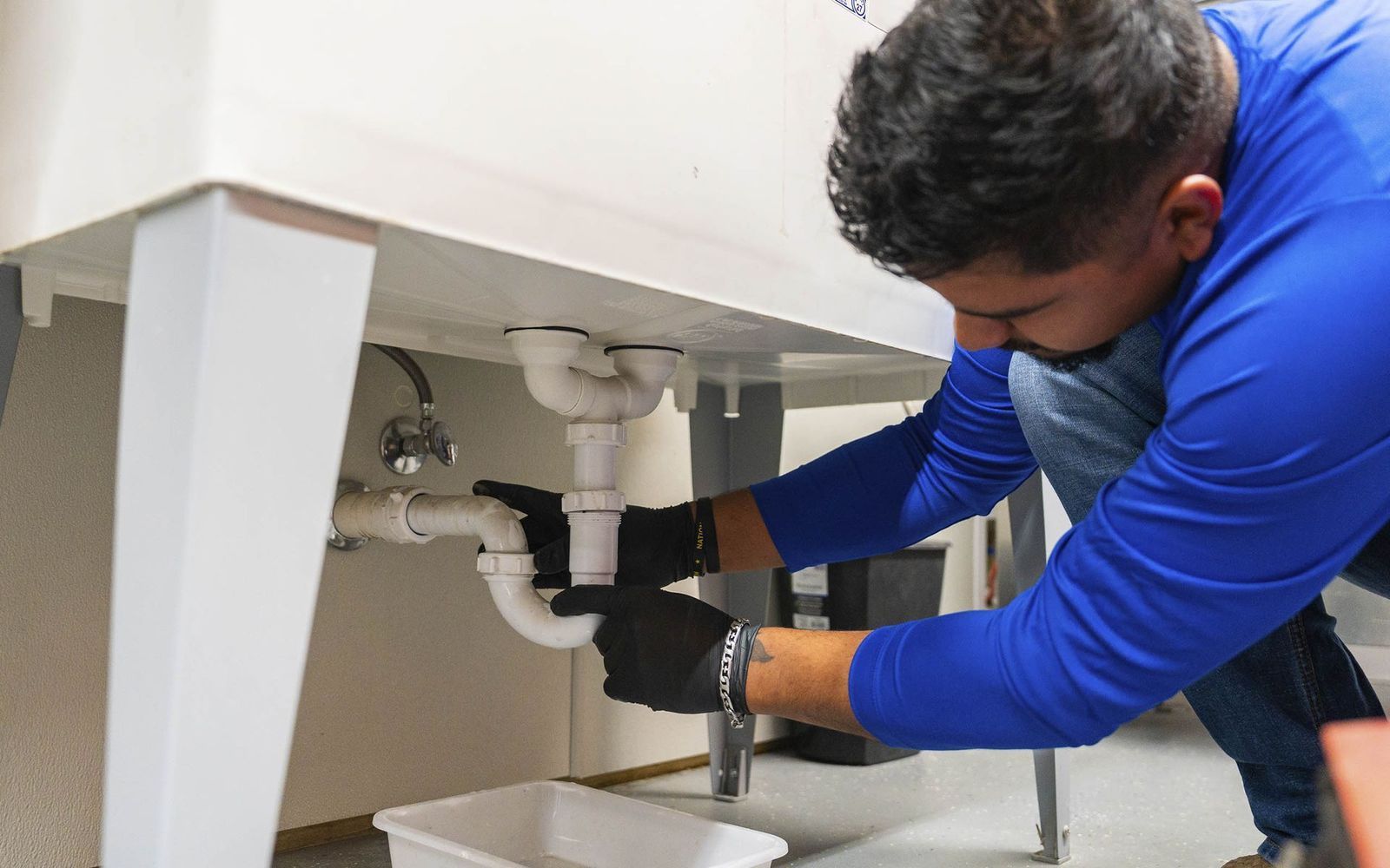 A man wearing gloves works on plumbing under a white sink.