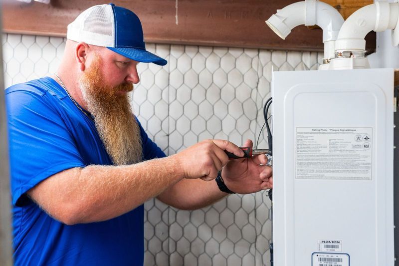 A bearded man in blue shirt and cap installs a white appliance, inside a room.