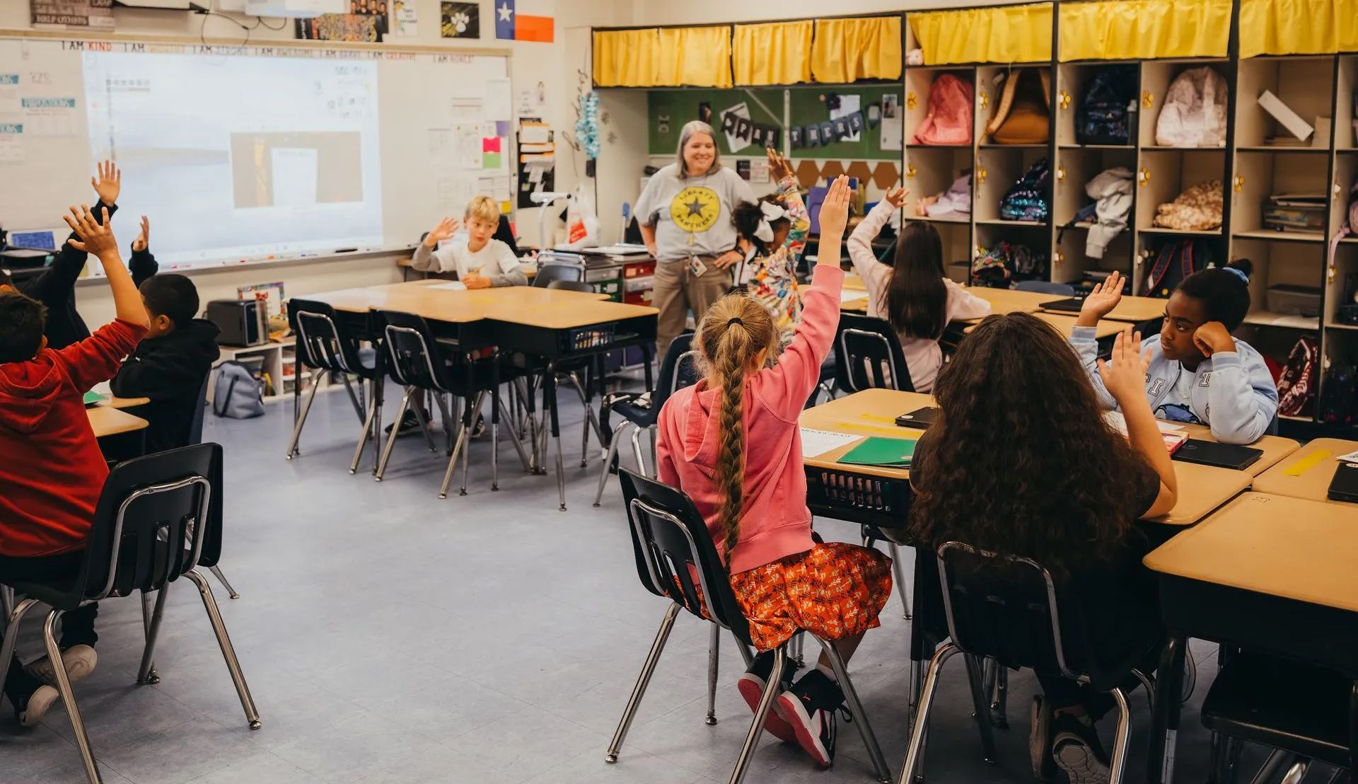 Students seated in a classroom, one child raising their hand to speak to their teacher