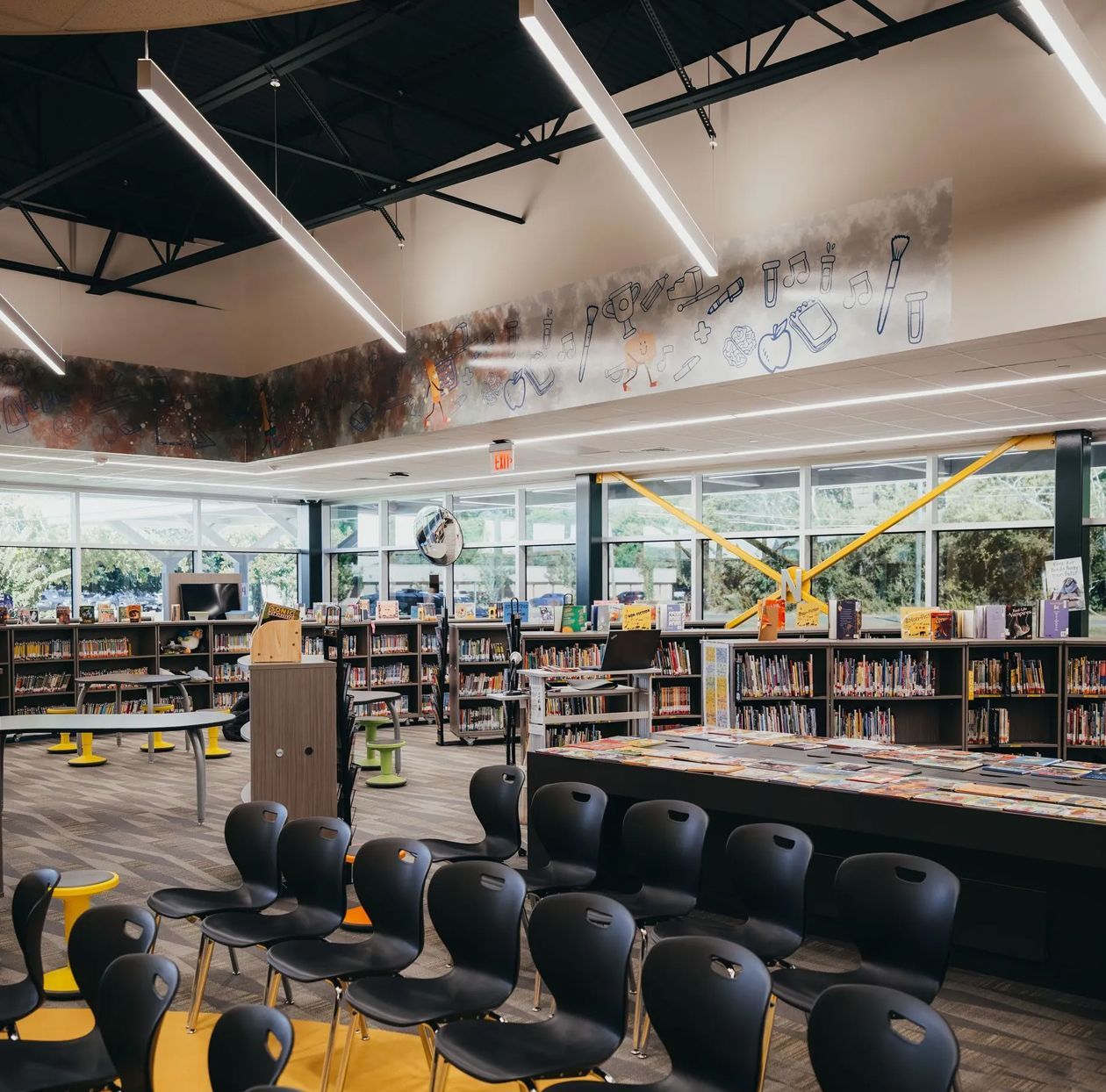 Library interior with rows of books, tables, and chairs, with large windows and a mural.