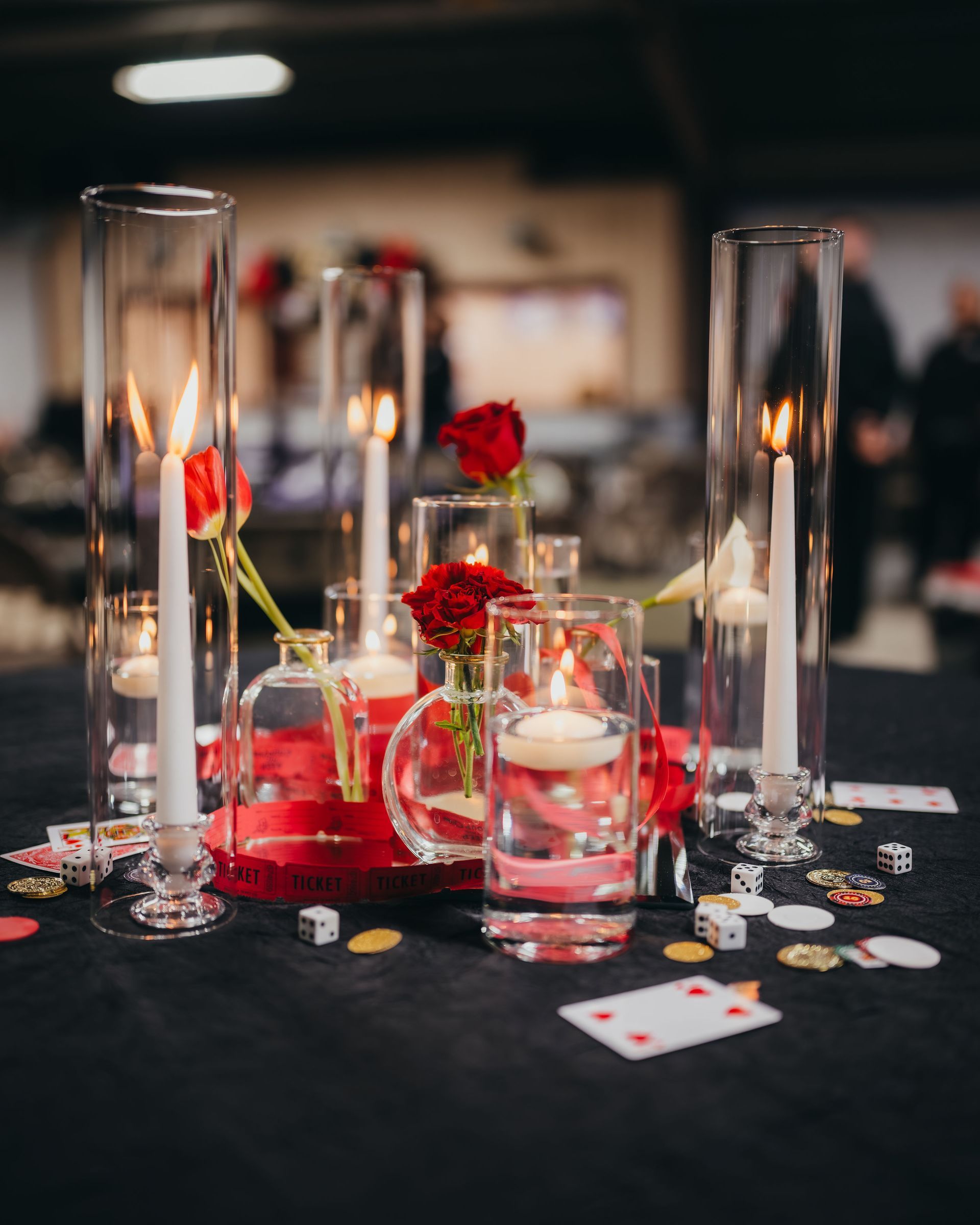 Centerpiece with red roses, candles in glass vases, and playing card accents on a black tablecloth.