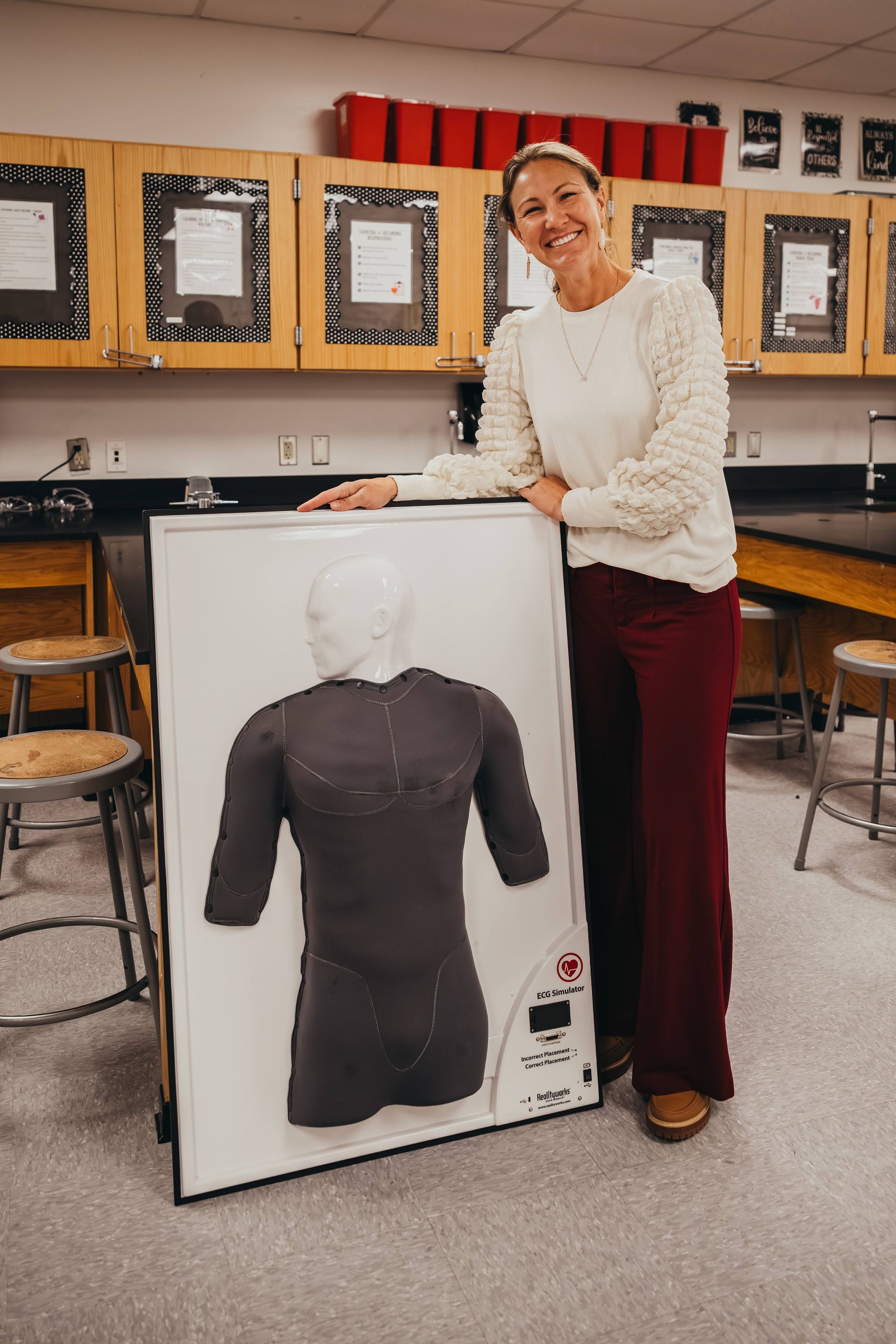 Woman in a classroom, smiling, stands next to a torso mannequin within a framed display.