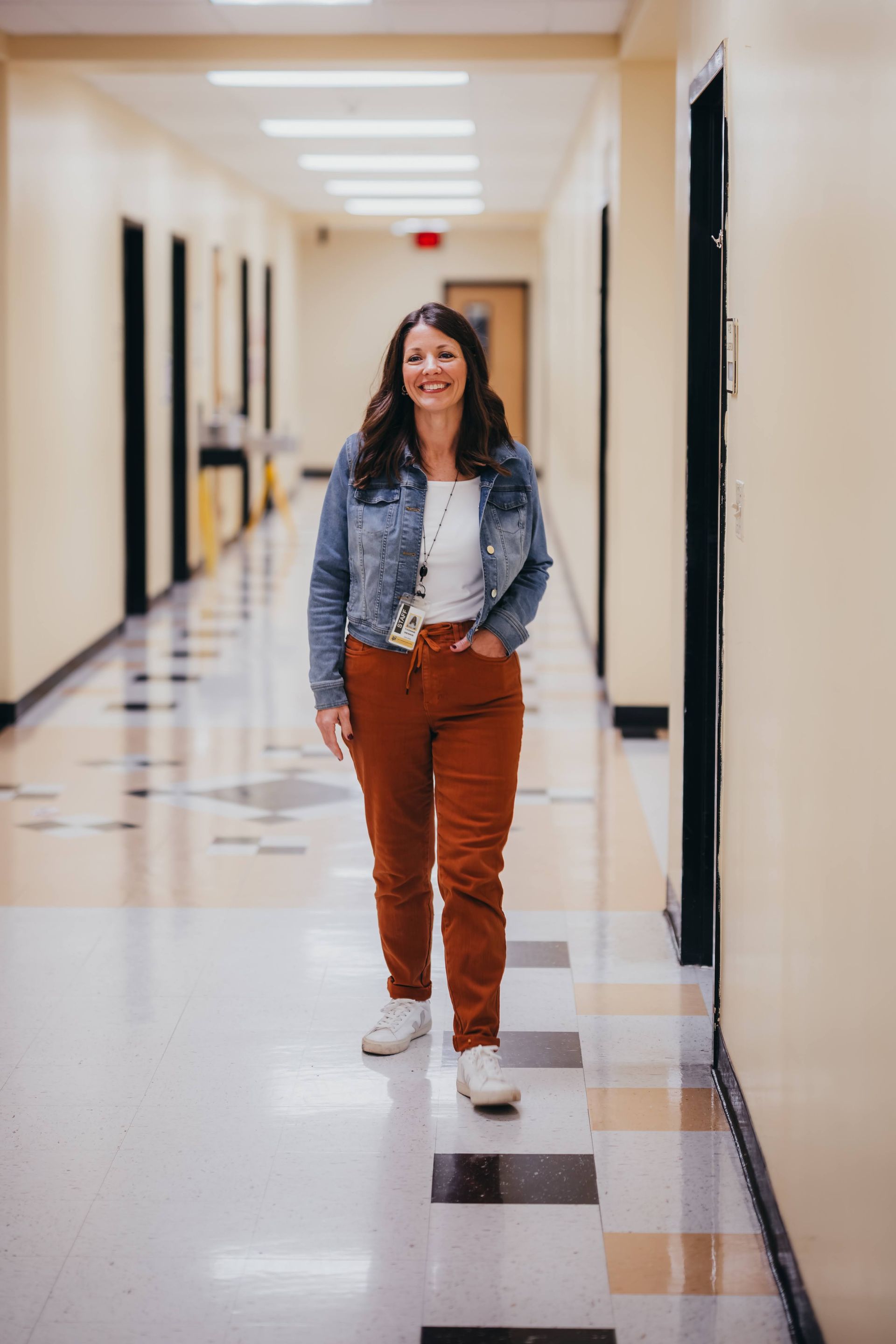 Woman in denim jacket, orange pants, and white shoes walks down a school hallway, smiling.