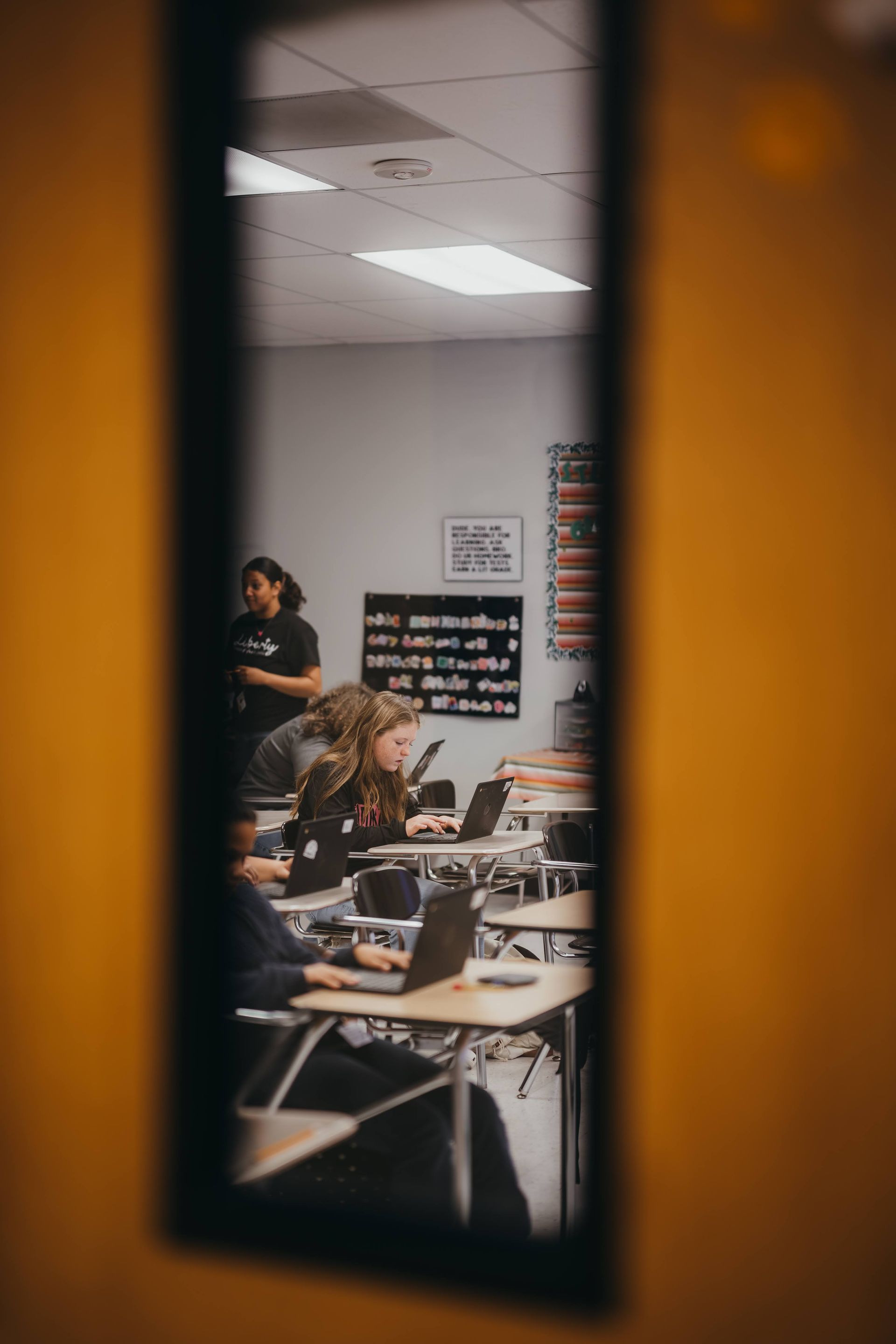 View of a classroom through a door window. Students work on laptops, and a person stands at the front.