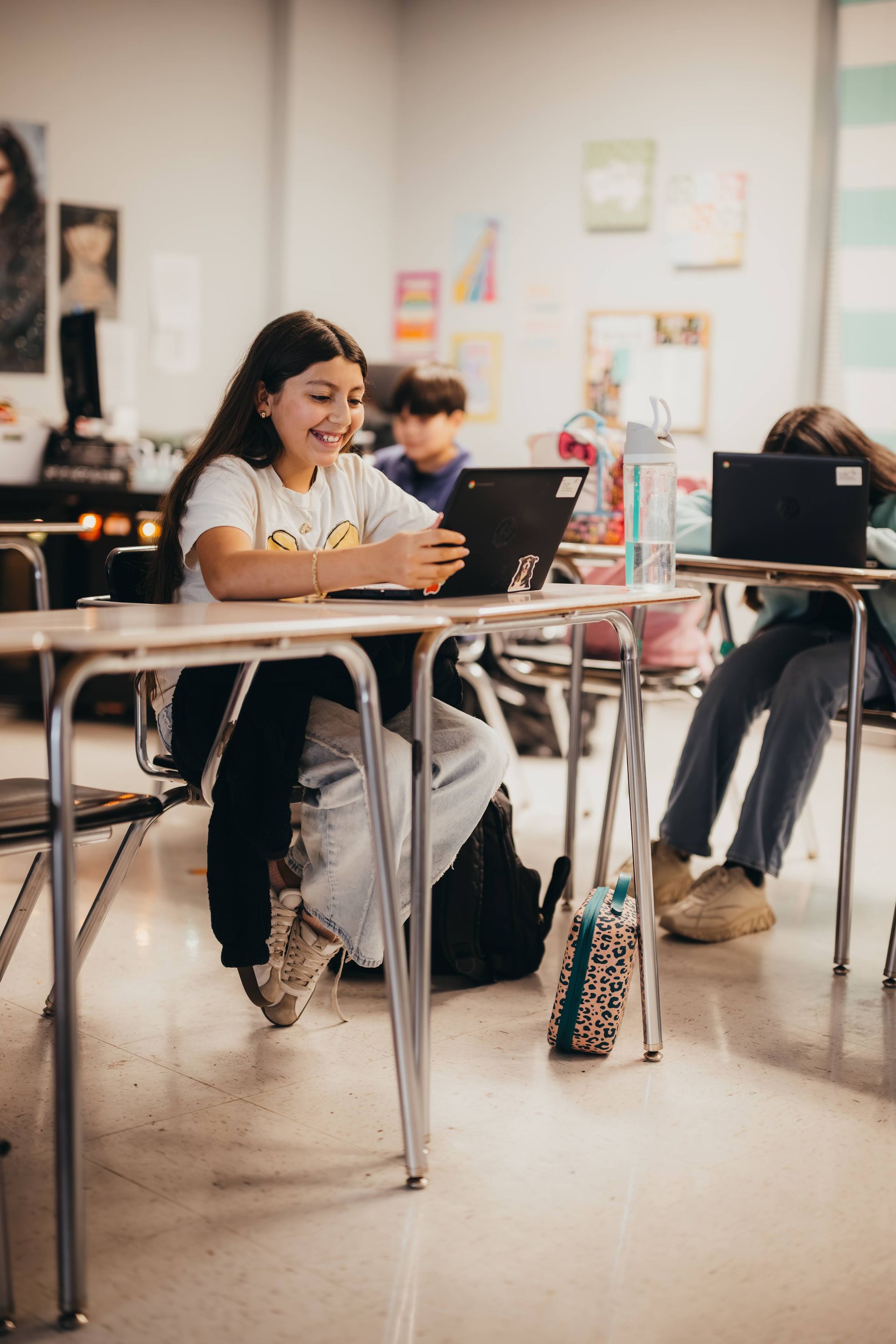 Girl smiles while using a tablet at a desk in a classroom; other students and desks visible.