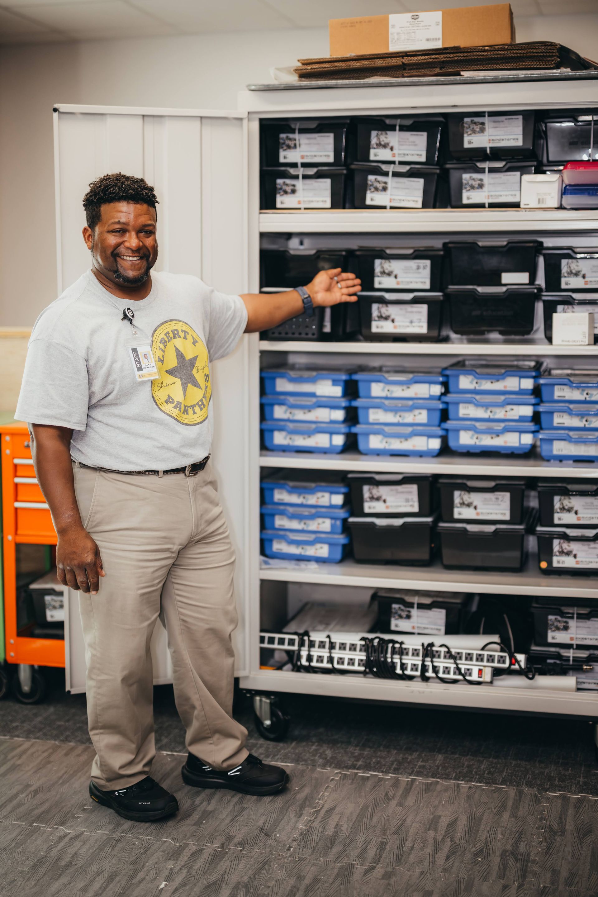 Man points to shelves filled with boxes, smiling. Light-colored pants, t-shirt. White shelves, orange tool cart.