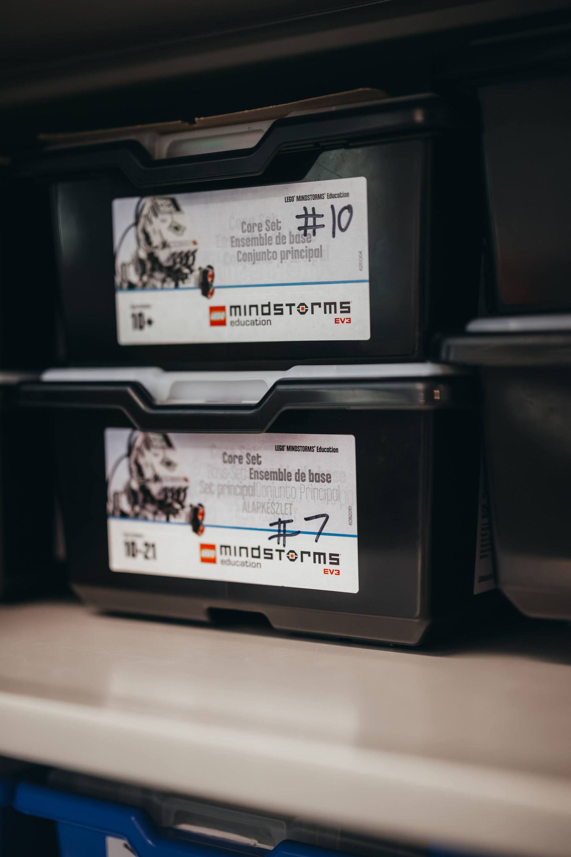 Two black LEGO MINDSTORMS storage containers on a shelf, labeled with handwritten text.